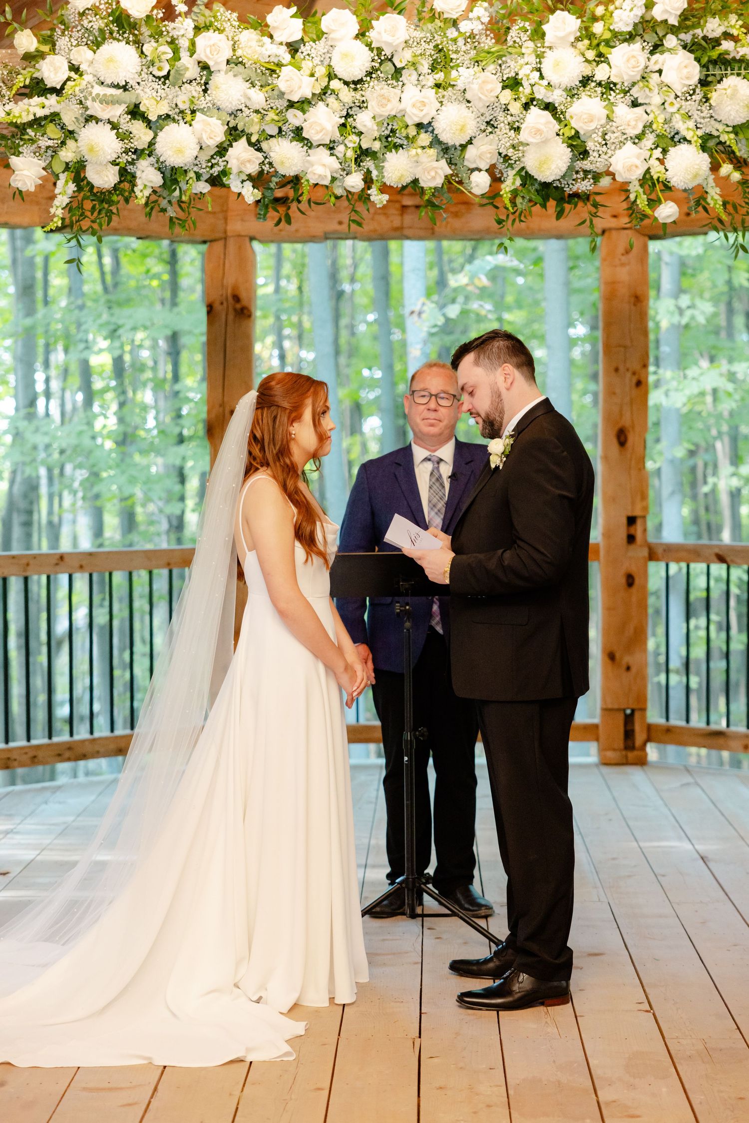 Wedding ceremony with elaborate white floral installation in rustic outdoor pavilion.