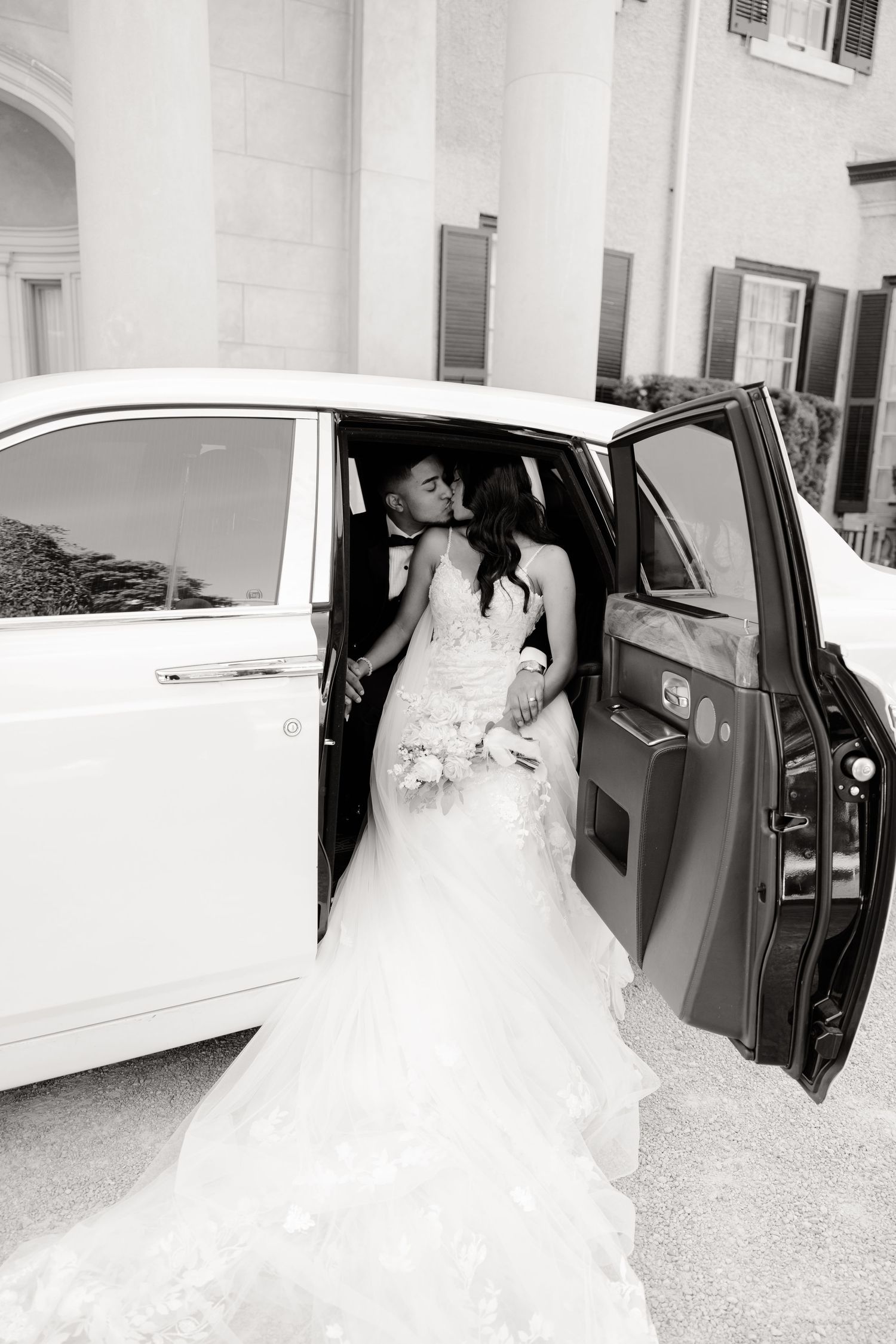 Black and white photo of newlyweds sharing a kiss in a vintage car.
