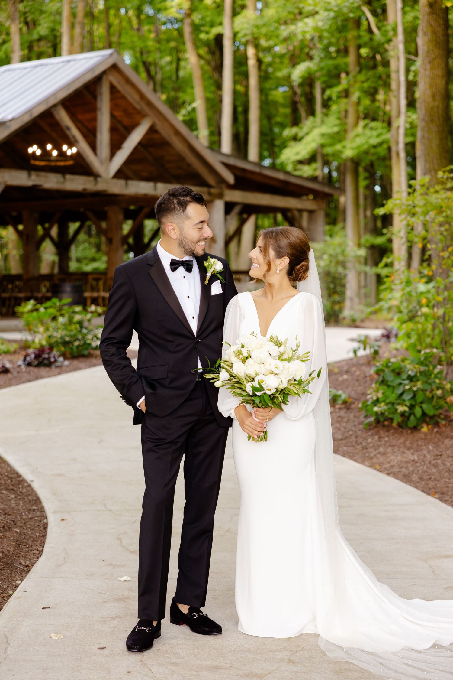 Bride and groom share intimate moment on curved garden path with rustic pavilion in background.