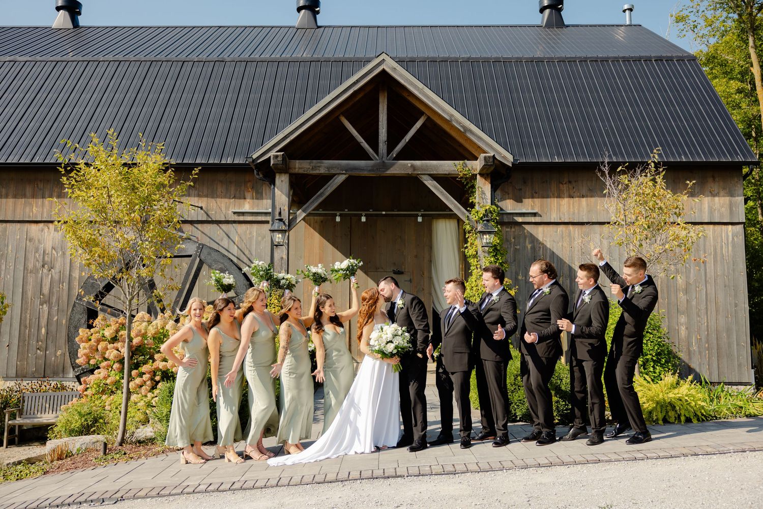 Wedding party celebrates with raised arms in front of a rustic barn with metal roof.