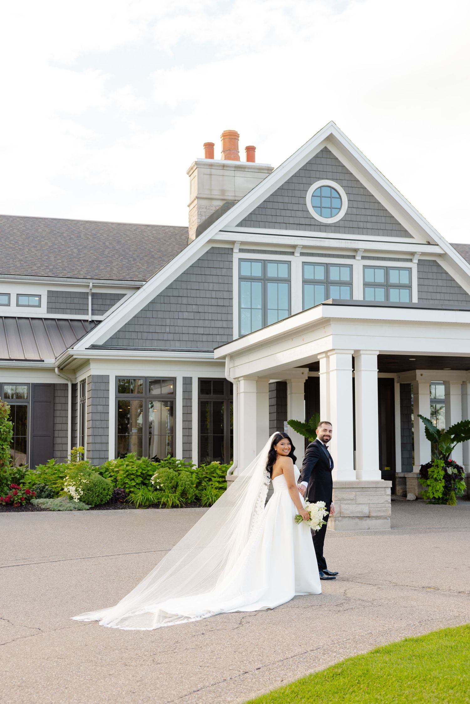 A couple stands outside an elegant gray house on their wedding day, with a flowing veil trailing behind.