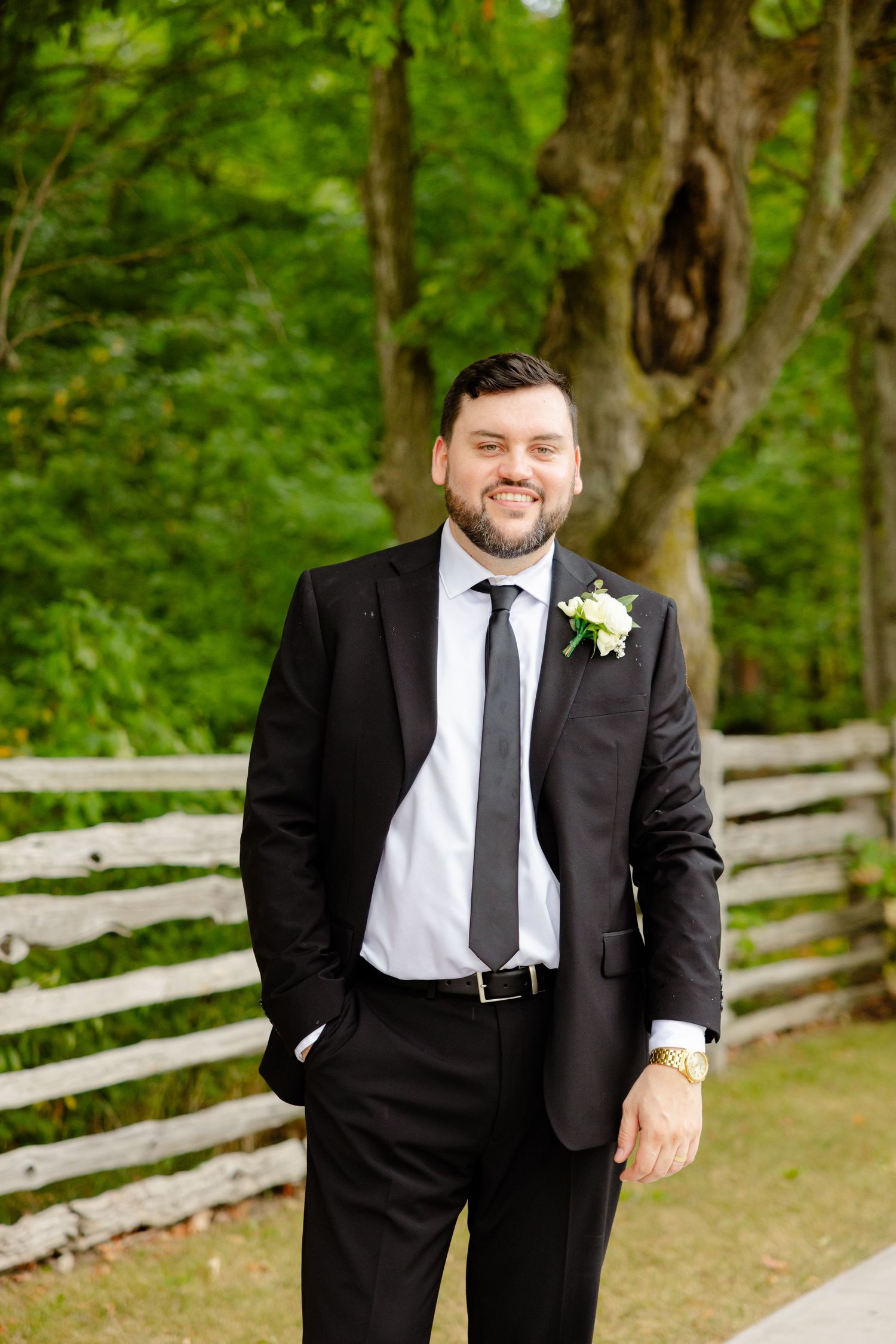Groom in classic black suit poses by rustic fence outdoors.