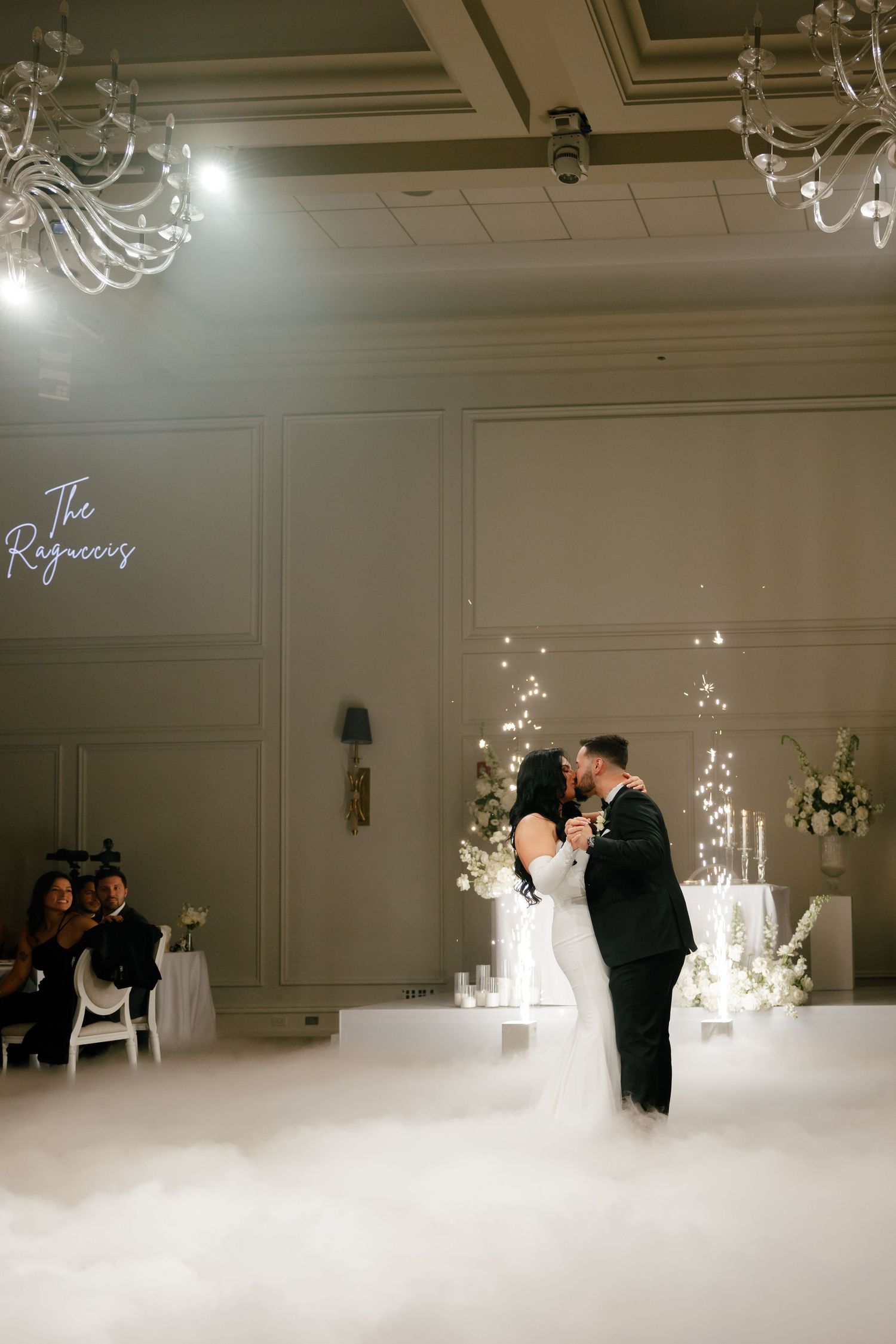 A couple shares their first dance in an elegant ballroom with fog effects and crystal chandeliers overhead.