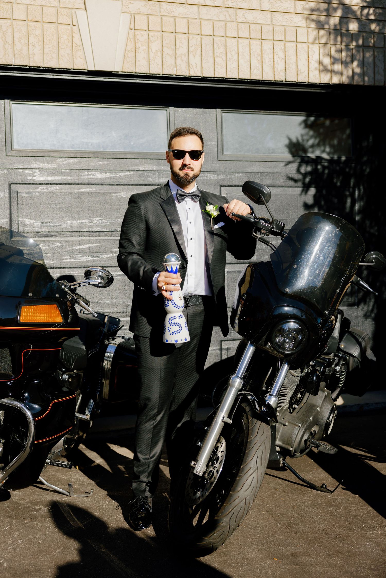A suited figure holds a drink while posing with motorcycles outside a garage.