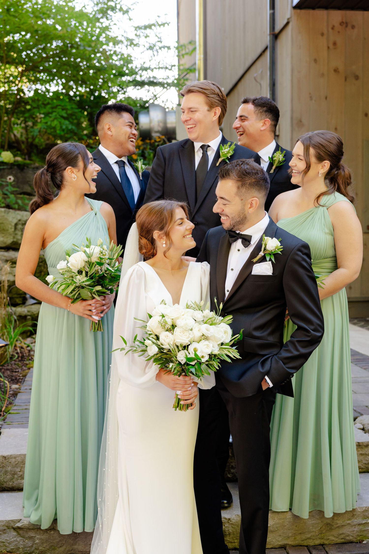 Wedding party members gather closely for casual photo outside rustic venue.