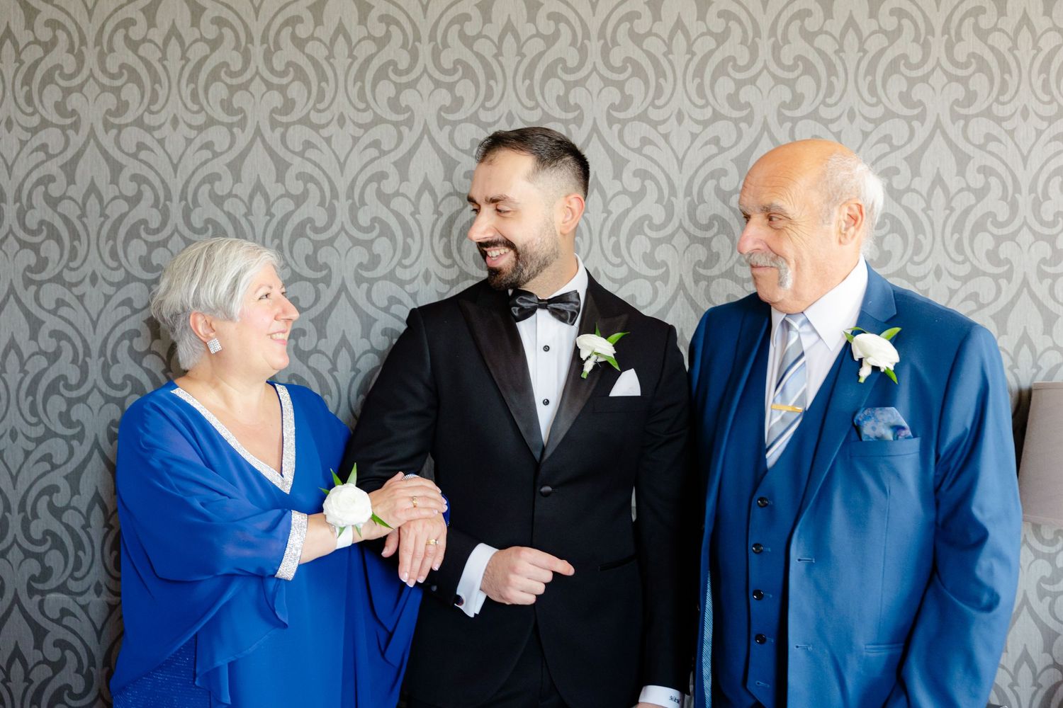 Three people in formal wear sharing a lighthearted moment against a grey patterned wall.