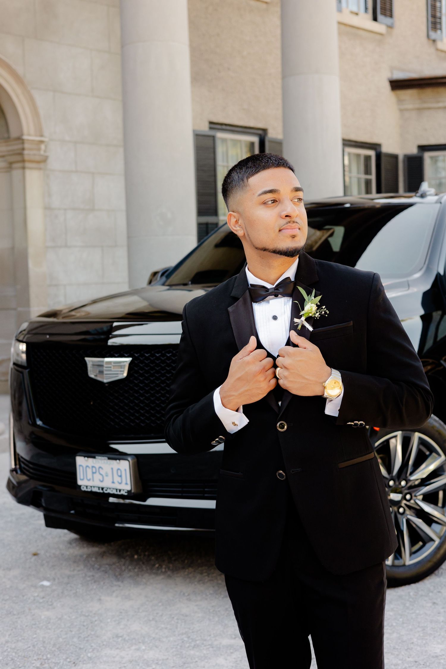 Groom in formal tuxedo adjusts his jacket while posing with a black Cadillac SUV.