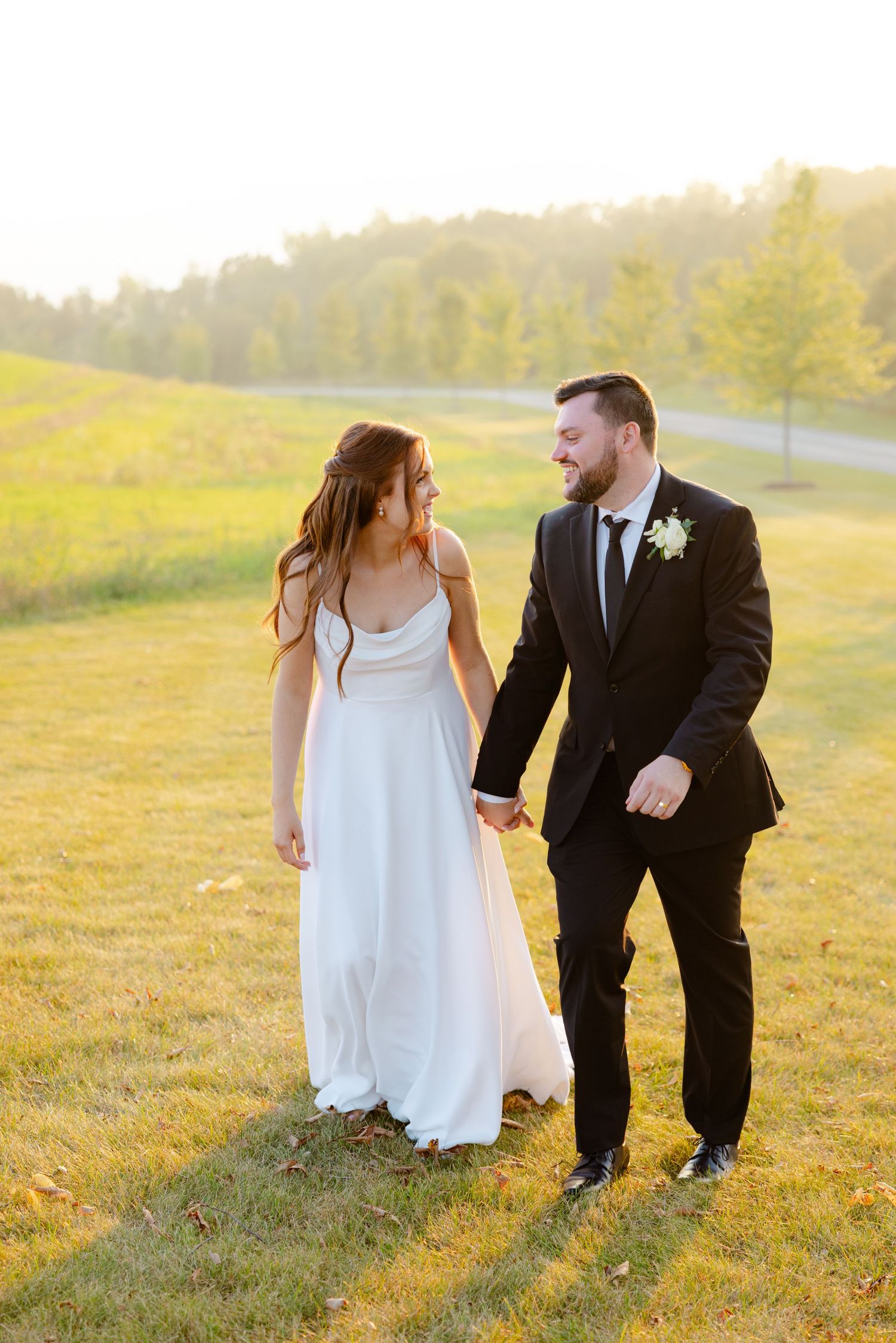 Wedding couple laughs together while walking on a grassy slope during golden hour.