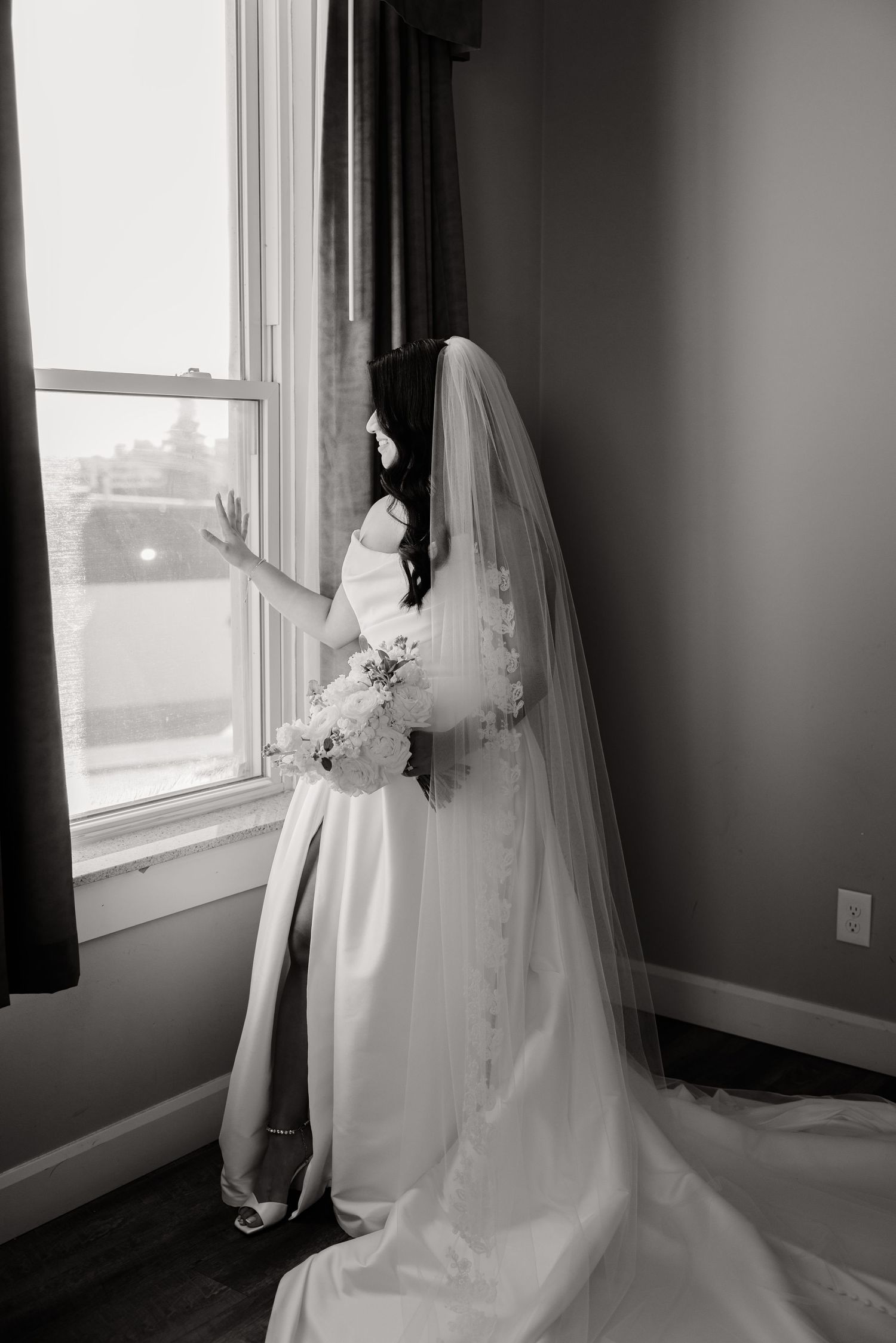 Black and white photo of bride in flowing dress and veil gazing out window.
