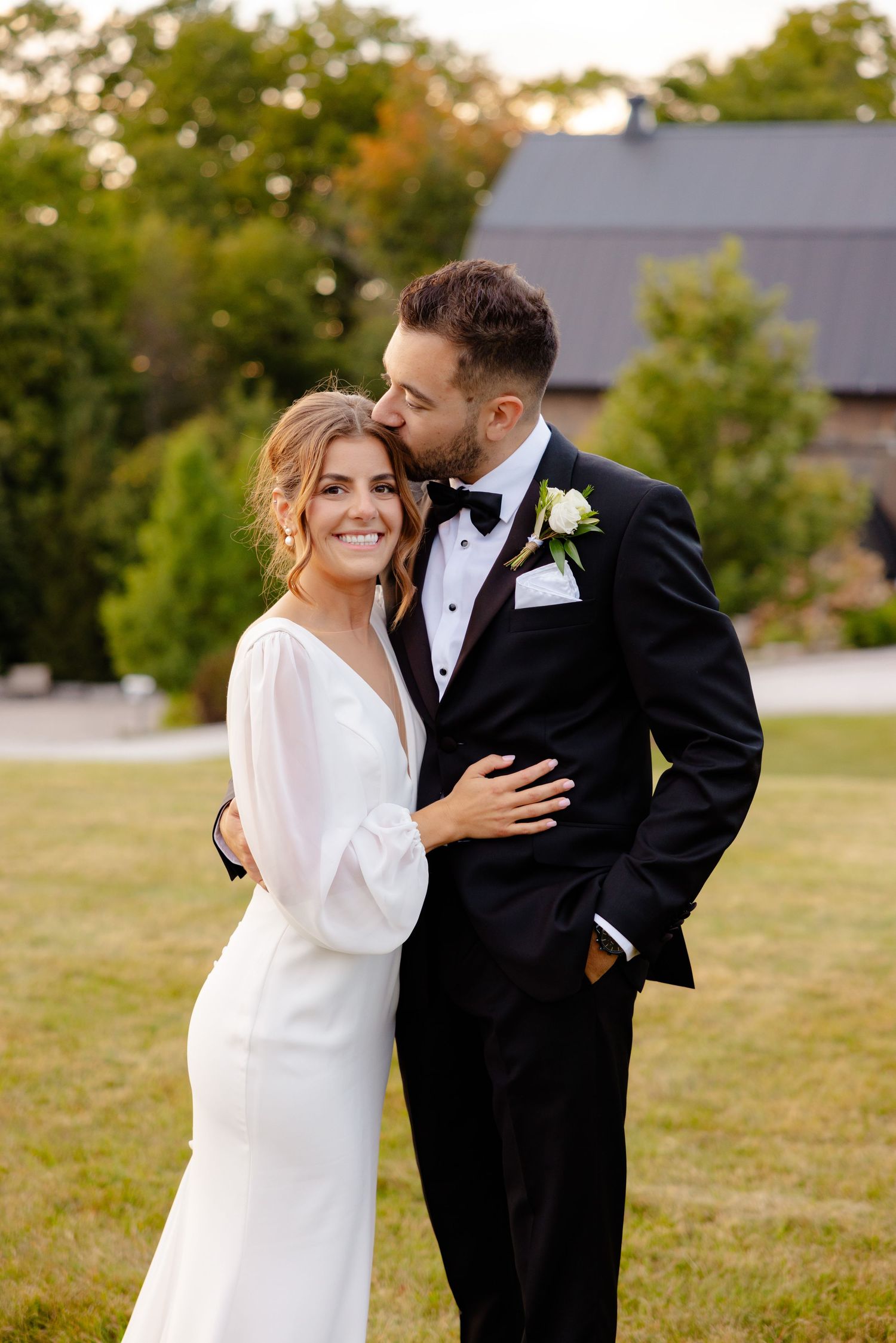 A romantic wedding portrait of a couple embracing outdoors with a barn in the background.
