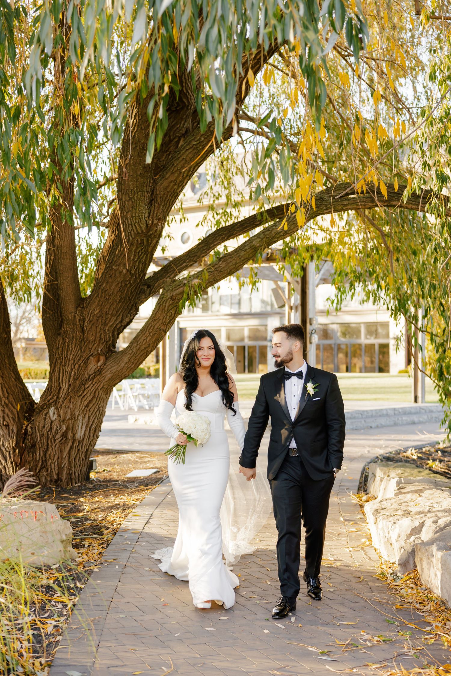 A couple in wedding attire walking hand in hand beneath a willow tree on a stone path.