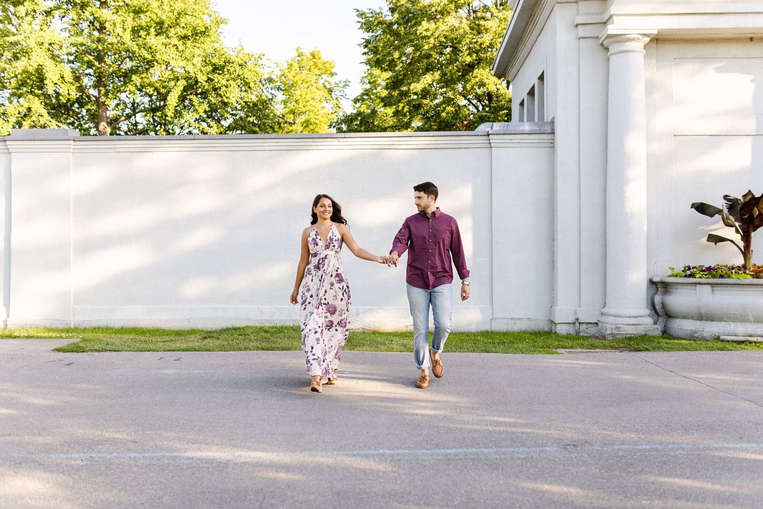 Couple walking hand in hand along white wall in burgundy shirt and floral dress.