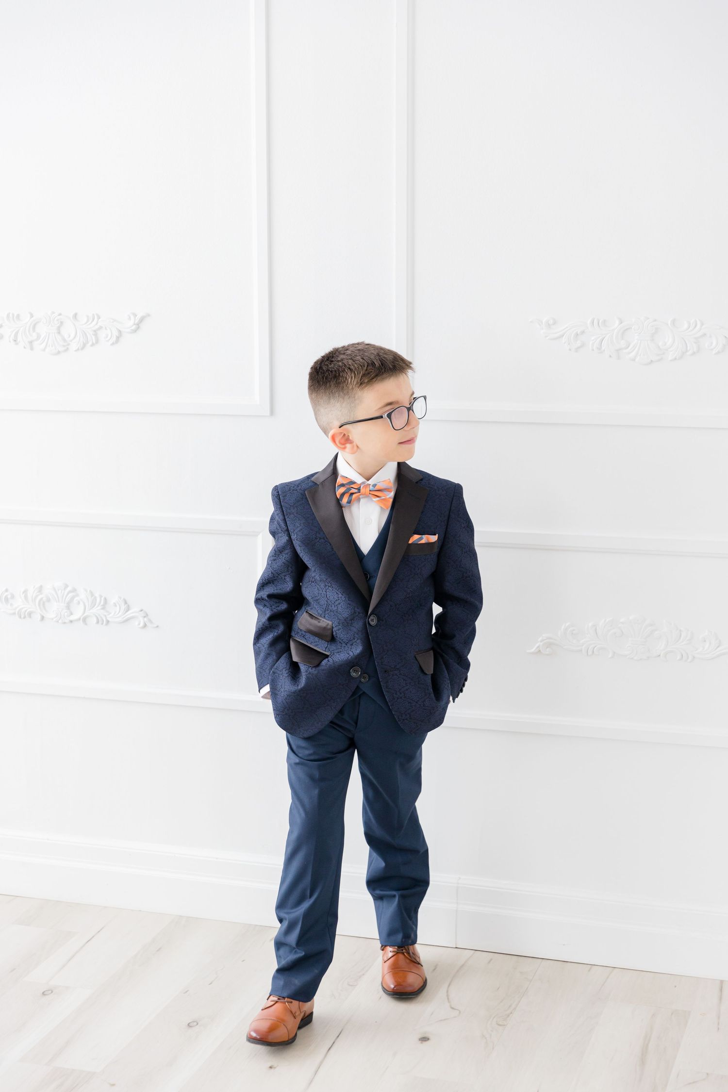 Youth in navy suit and glasses standing with hands in pockets against white backdrop.