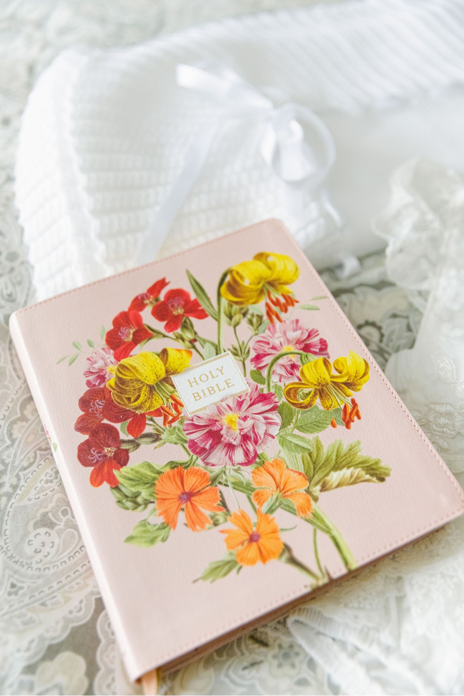 A pink book cover decorated with colorful floral illustrations resting on white lace fabric.