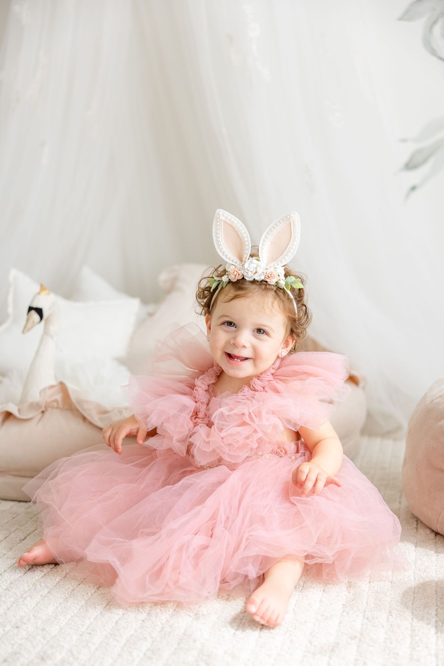 A toddler wearing a pink tulle dress and bunny ear crown sits on a light-colored floor.