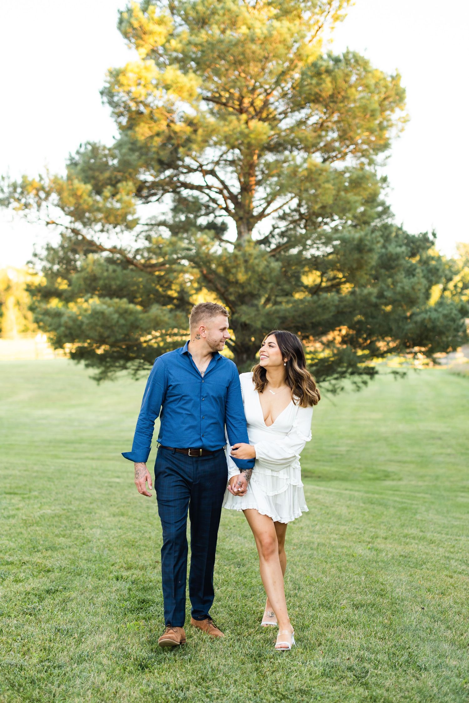 Couple strolling on green lawn with tall pine tree in background during sunset.
