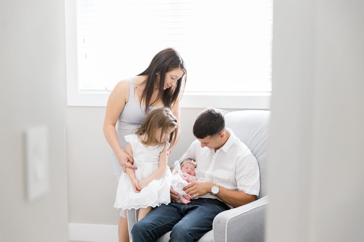 A family shares a tender moment together on a grey armchair in a bright nursery room.