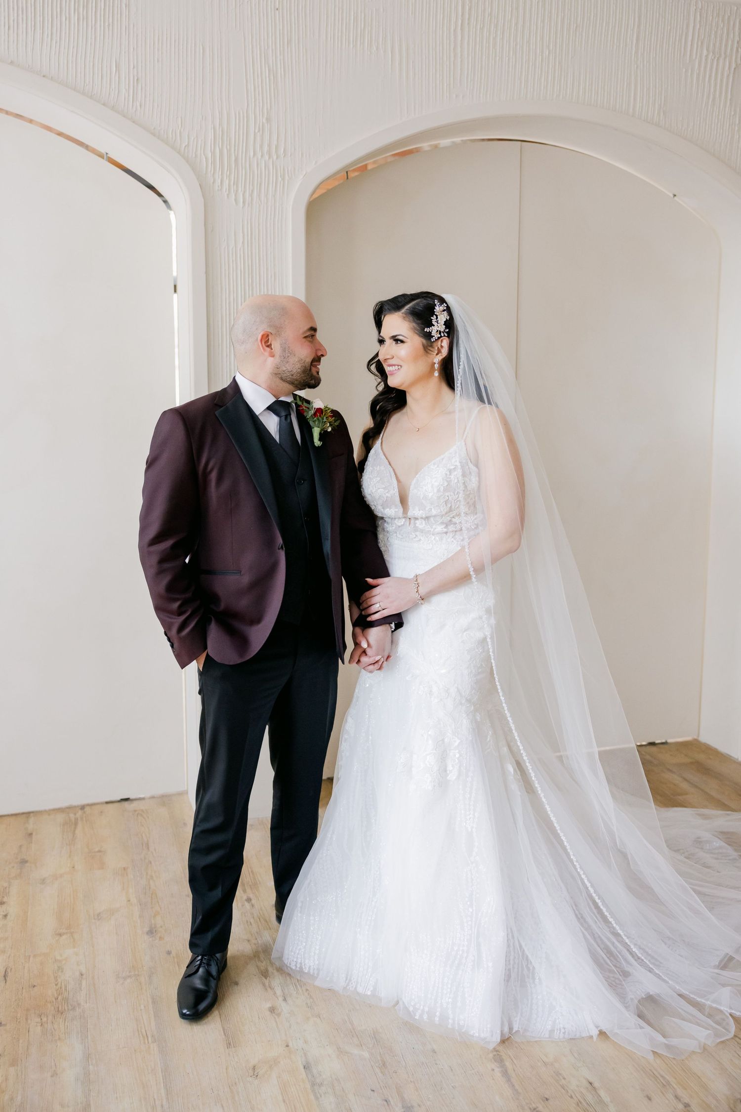 Elegant wedding portrait with groom in burgundy suit and bride in lace gown.