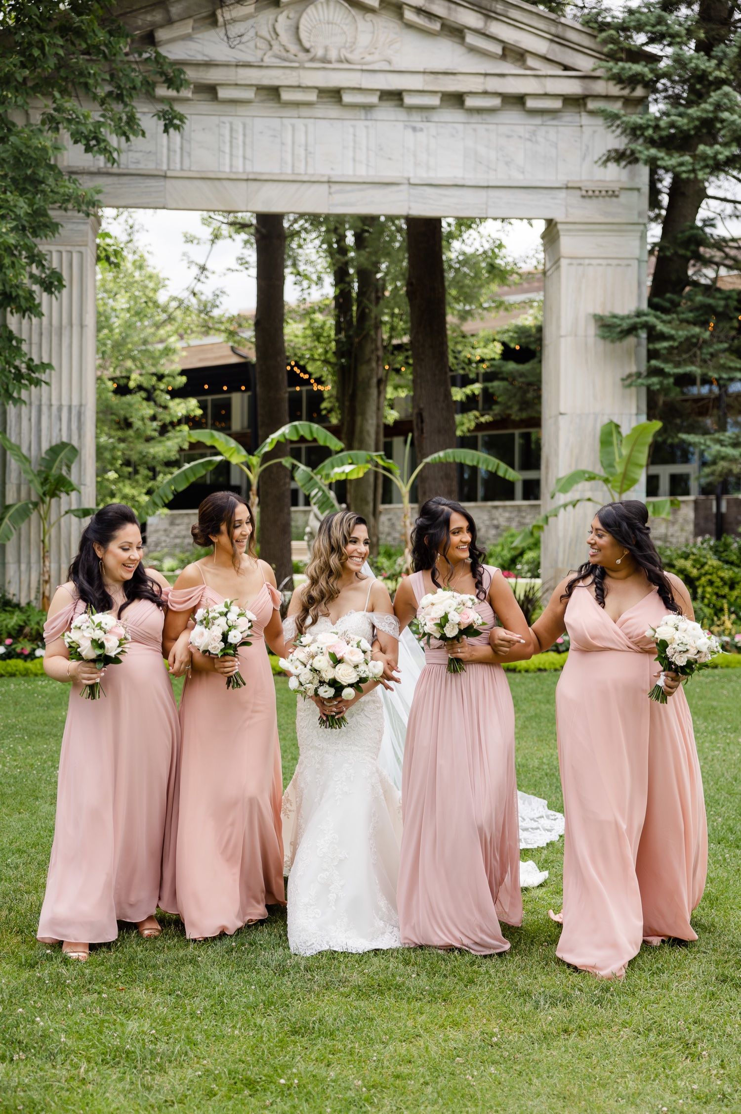 Bridal party wearing blush pink gowns poses informally under white stone archway outdoors.