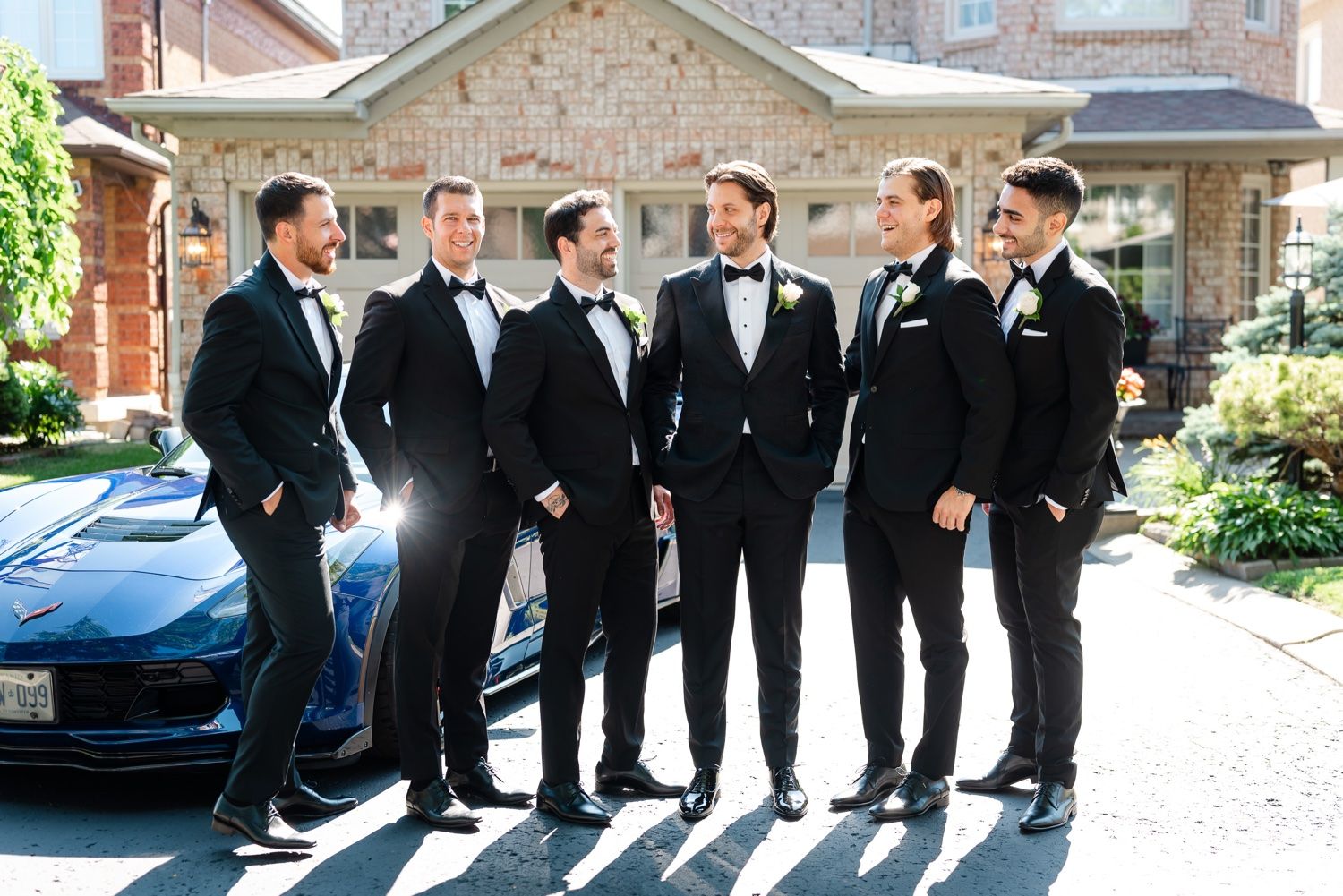 Group of groomsmen in black suits stand together outdoors in natural lighting.