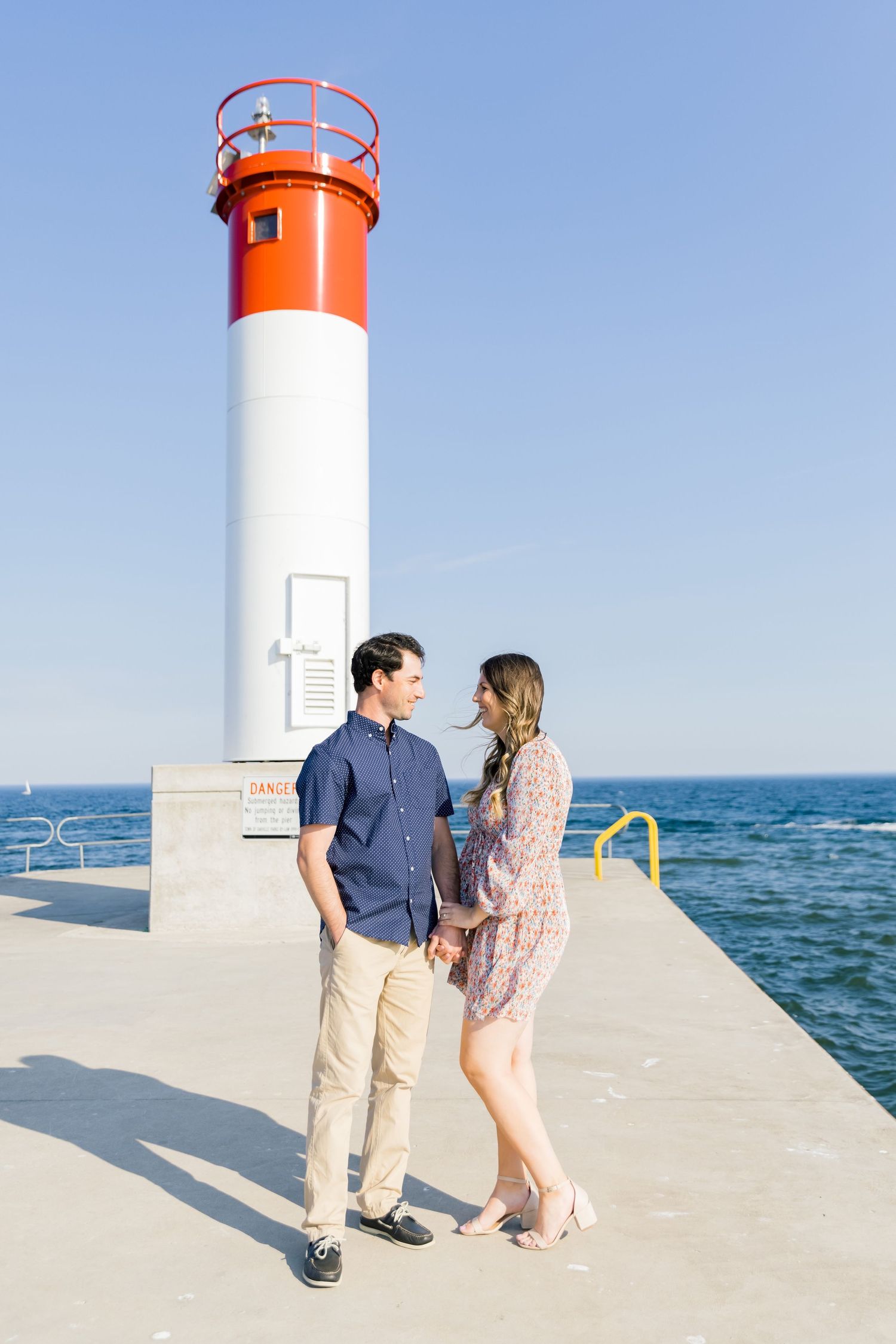 A couple stands together on a pier next to an orange and white lighthouse against a blue ocean backdrop.