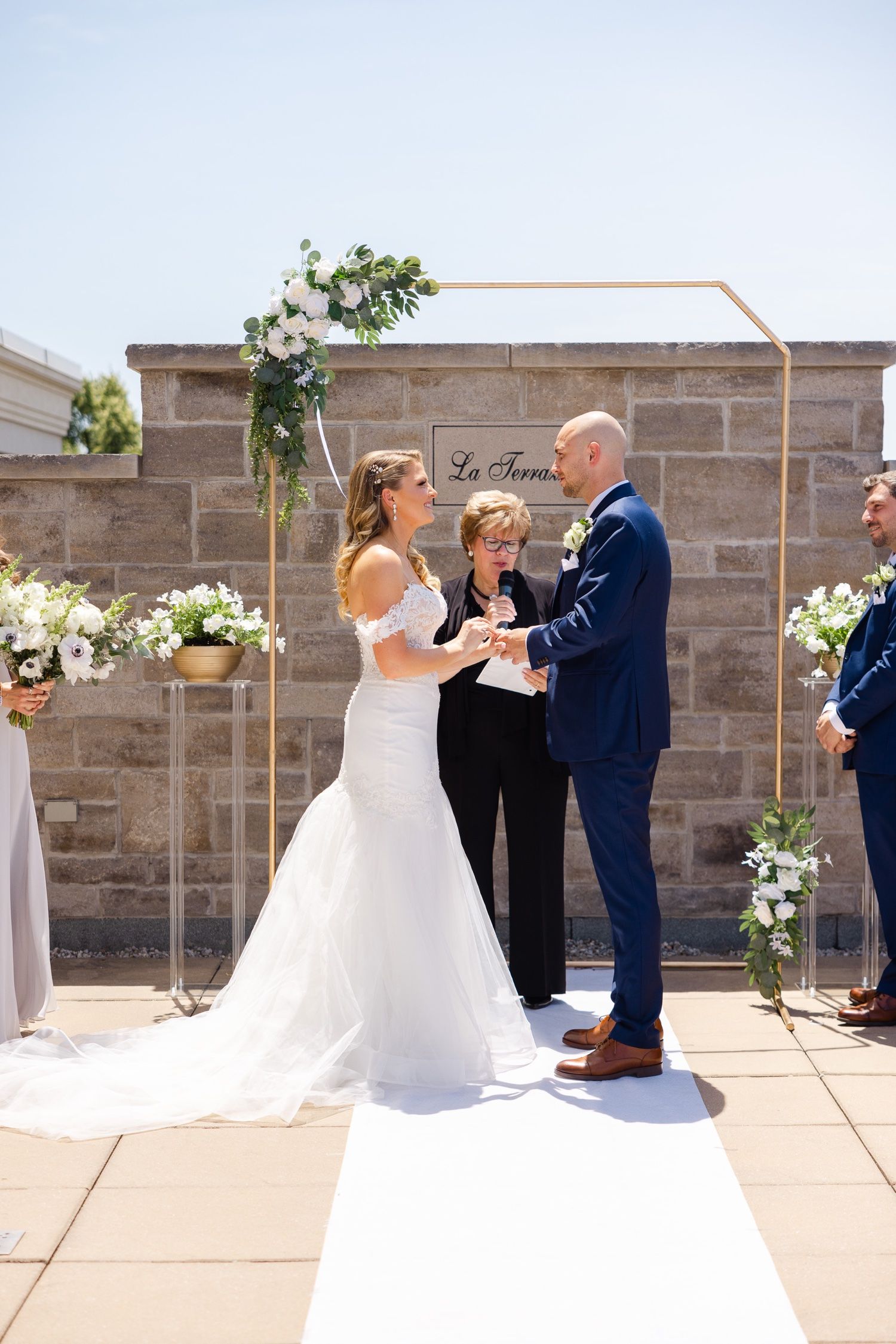 A wedding ceremony under a modern copper arch with white florals against a stone wall backdrop.