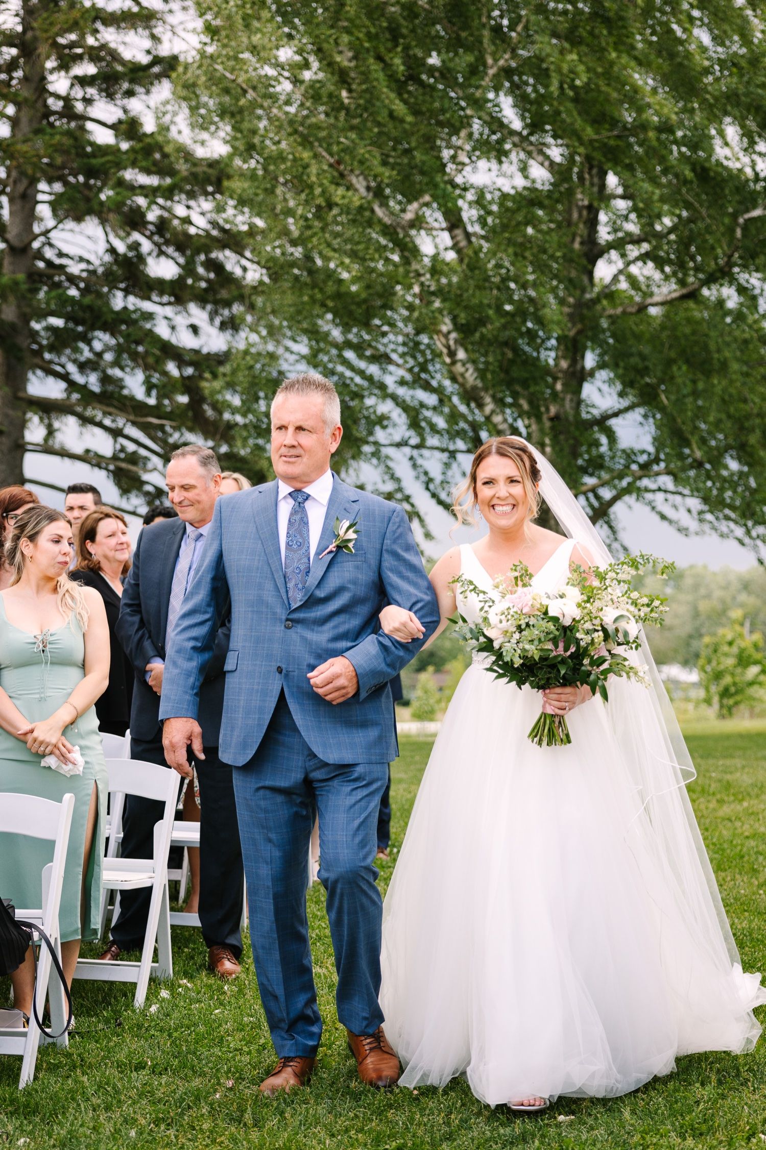 Wedding ceremony processional along aisle with guests seated outdoors under green trees.
