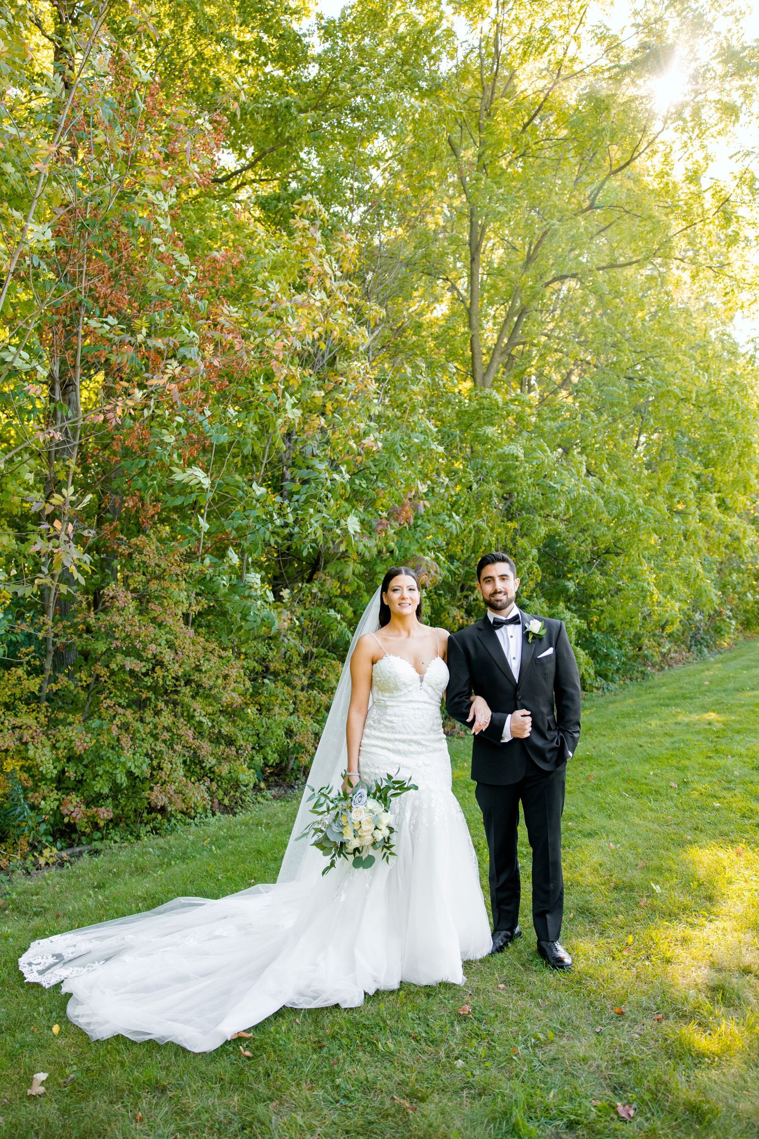 Couple poses for full-length wedding portrait on grassy hill with autumn foliage.