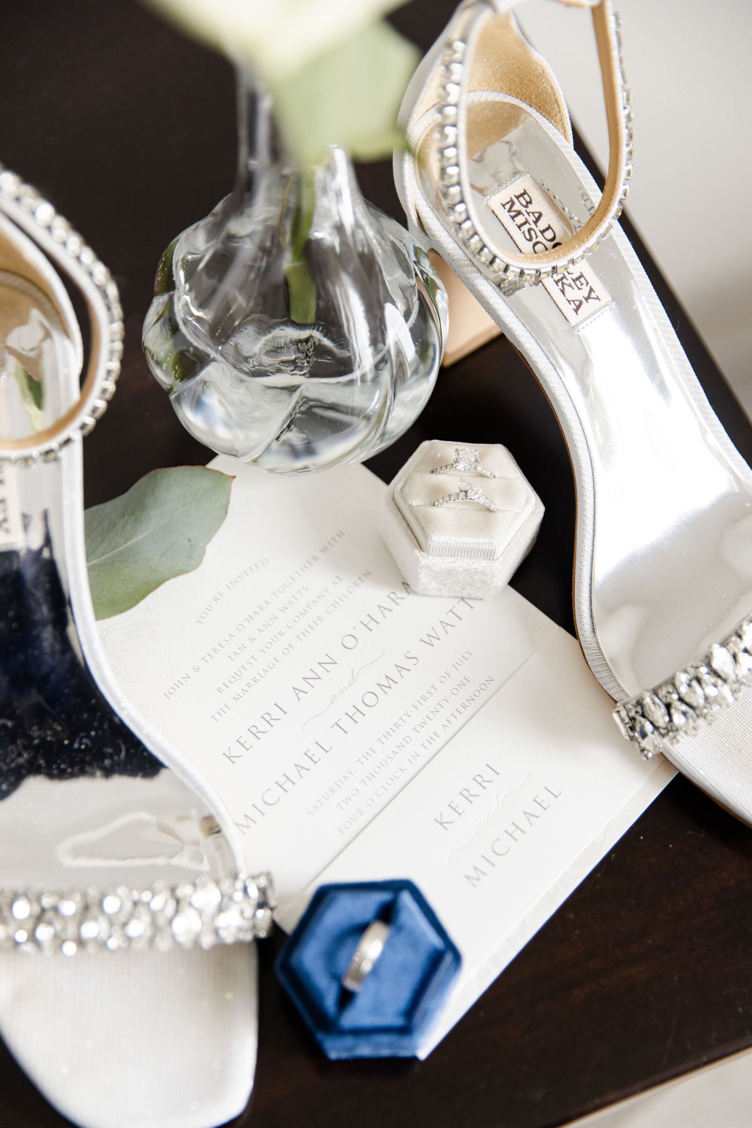 Wedding accessories including crystal-embellished shoes, ring boxes, and invitation suite on dark surface.
