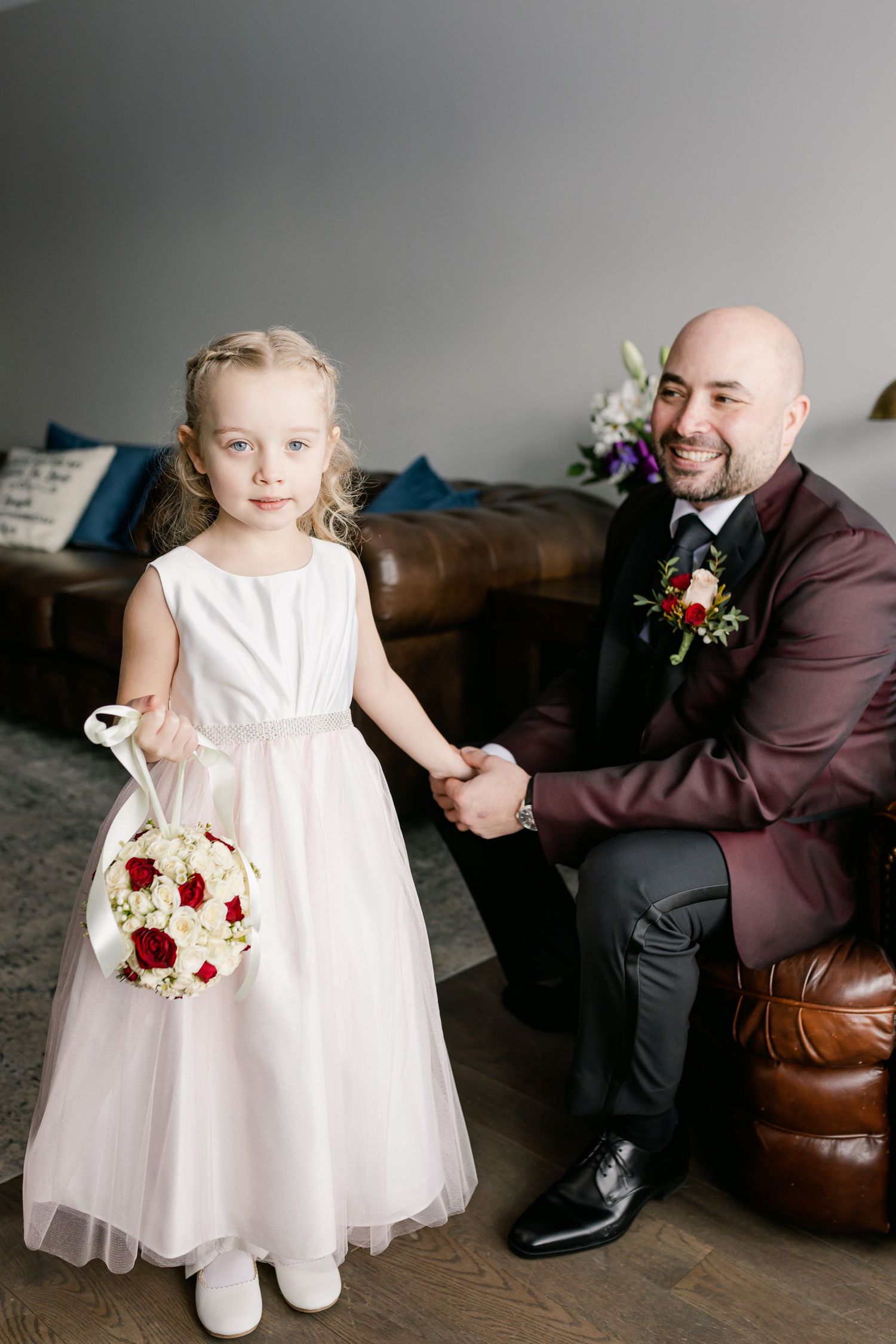 Flower girl in white dress holding red and white bouquet next to man in burgundy suit.