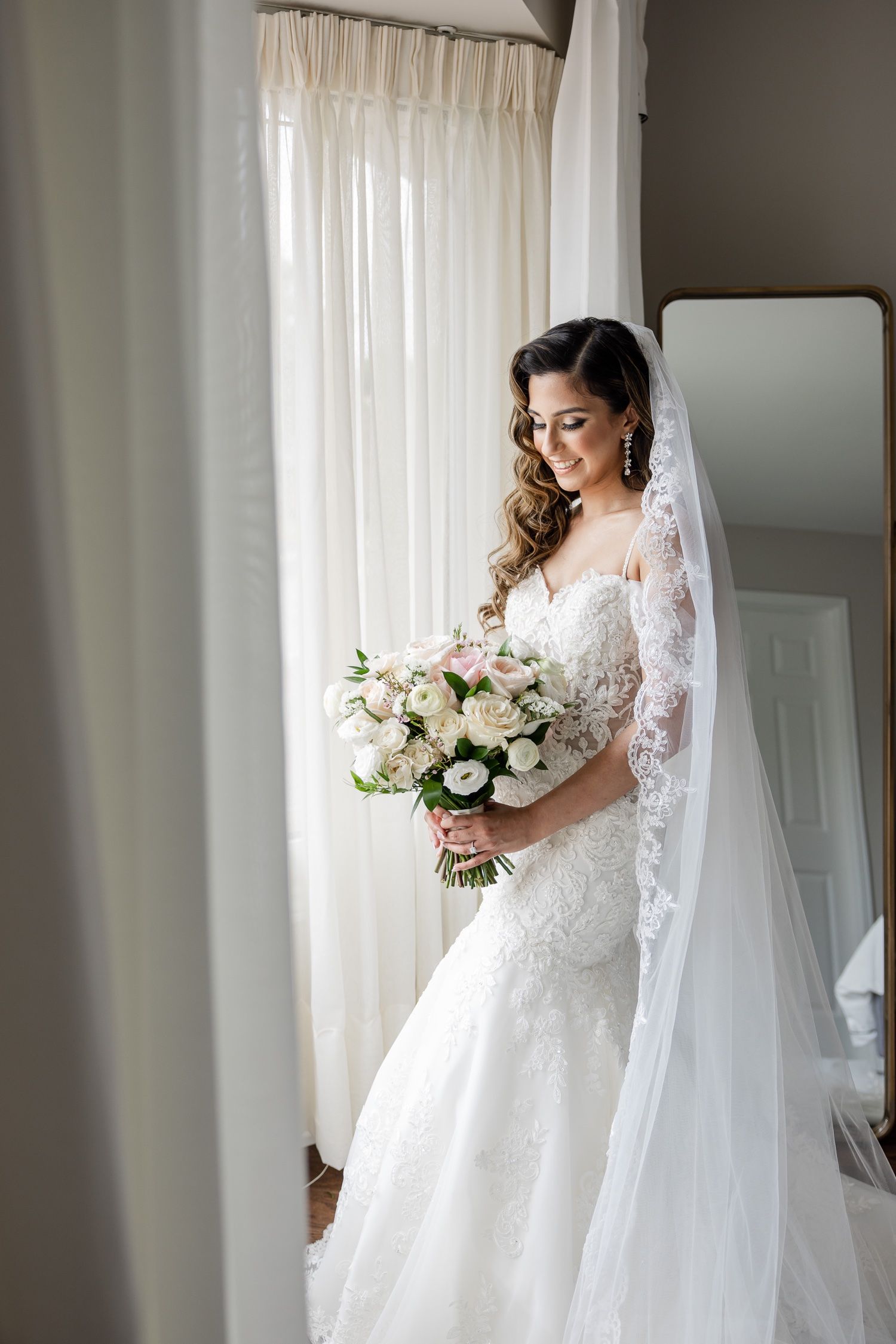 Bride in lace wedding gown holding white bouquet poses by window in natural light.