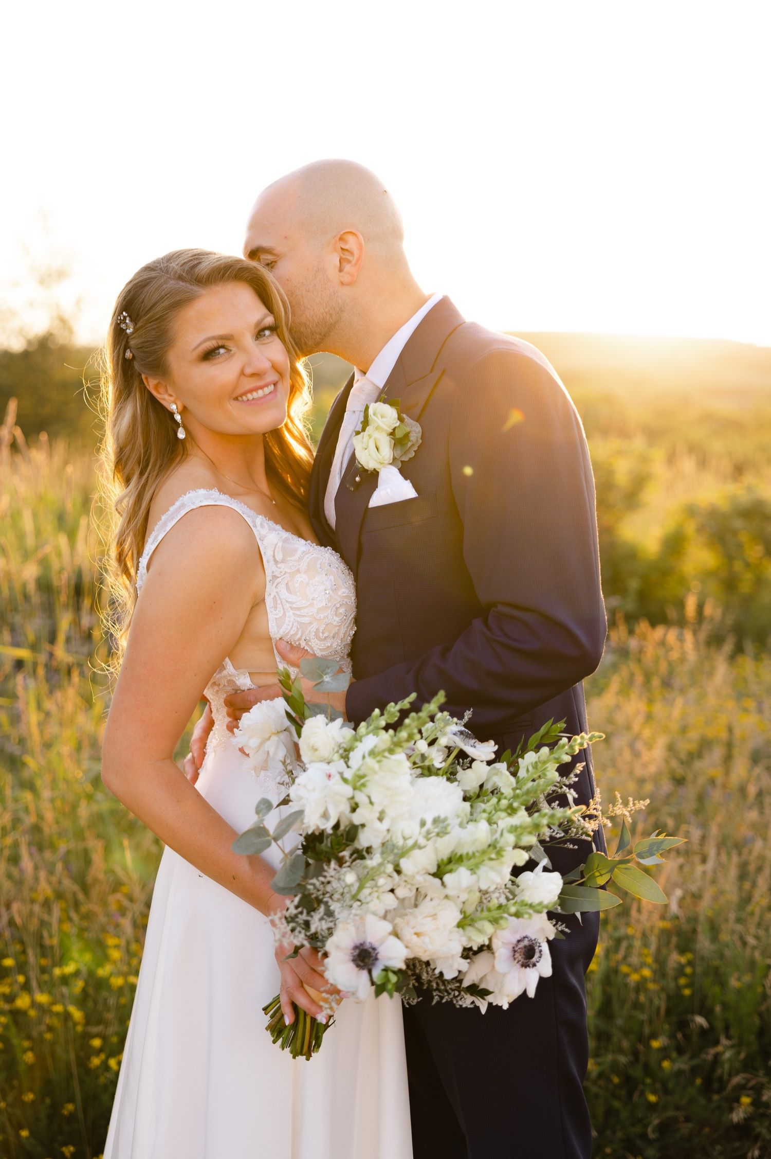 A wedding couple poses in a field at sunset with a stunning white floral bouquet.