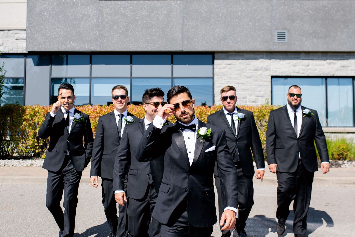 Sharp-dressed groomsmen in sunglasses walking confidently in front of modern building.