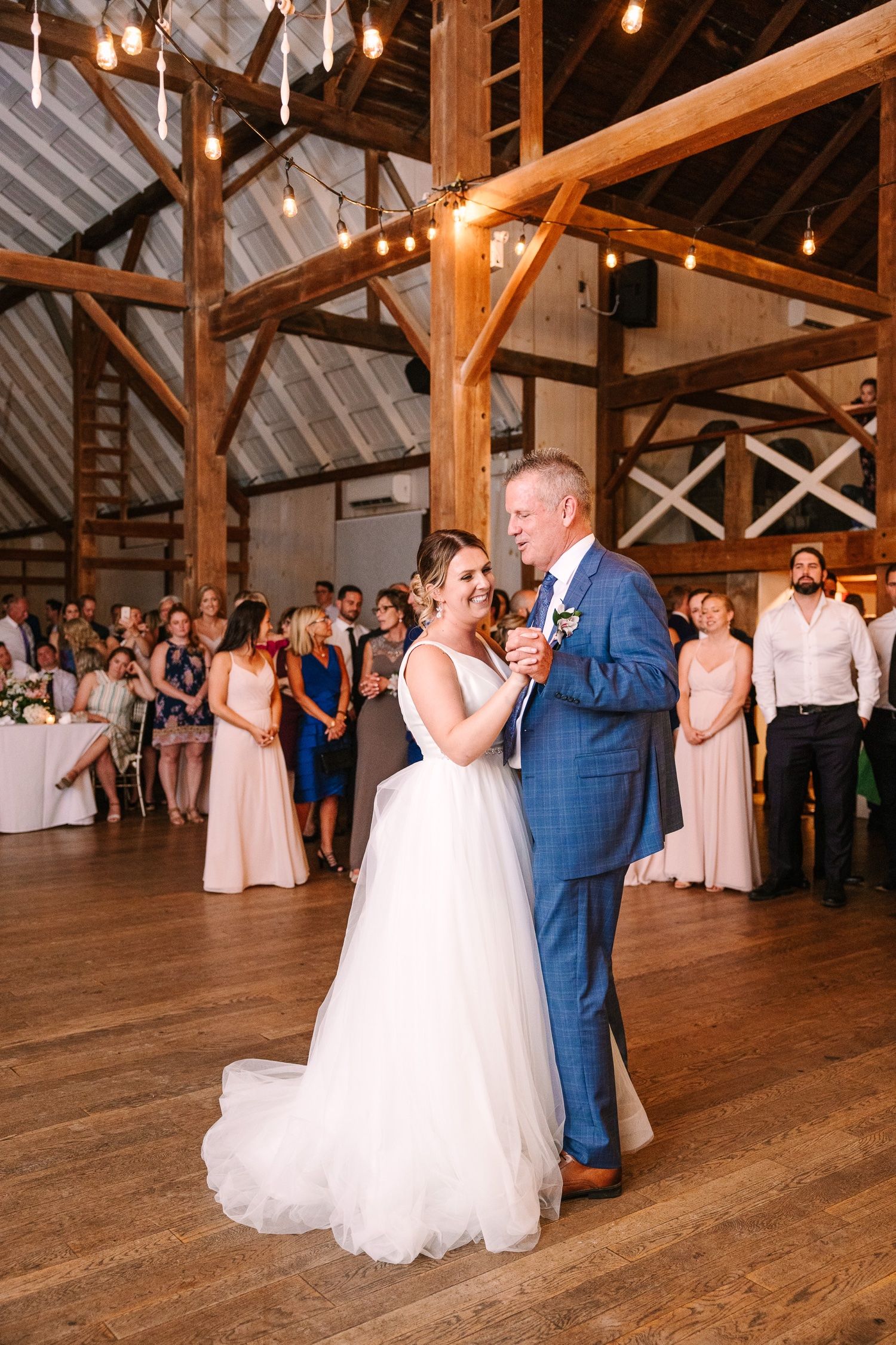 Newlyweds share an intimate dance in a rustic wood-beamed barn with wedding guests looking on.