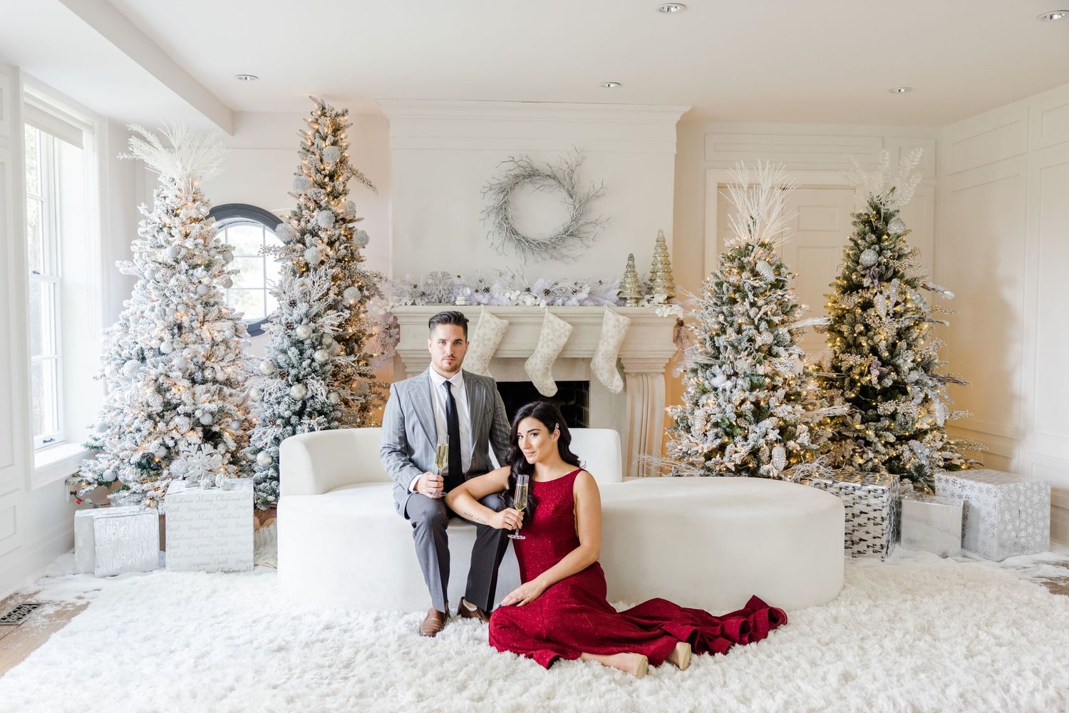 Elegant holiday photoshoot featuring couple in red dress and grey suit among decorated Christmas trees.