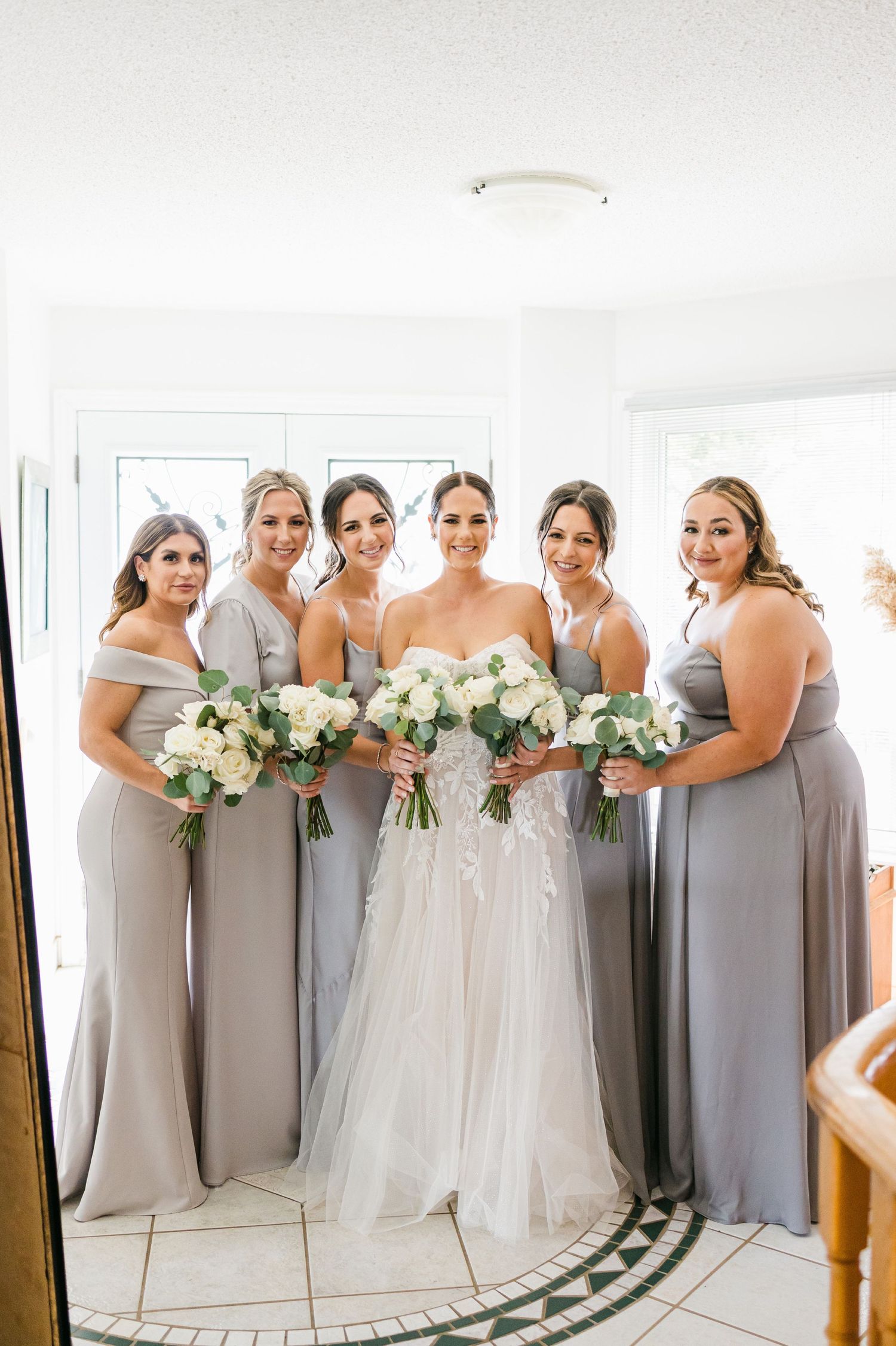 Bridesmaids in elegant grey dresses pose together with white floral bouquets in a bright interior space.