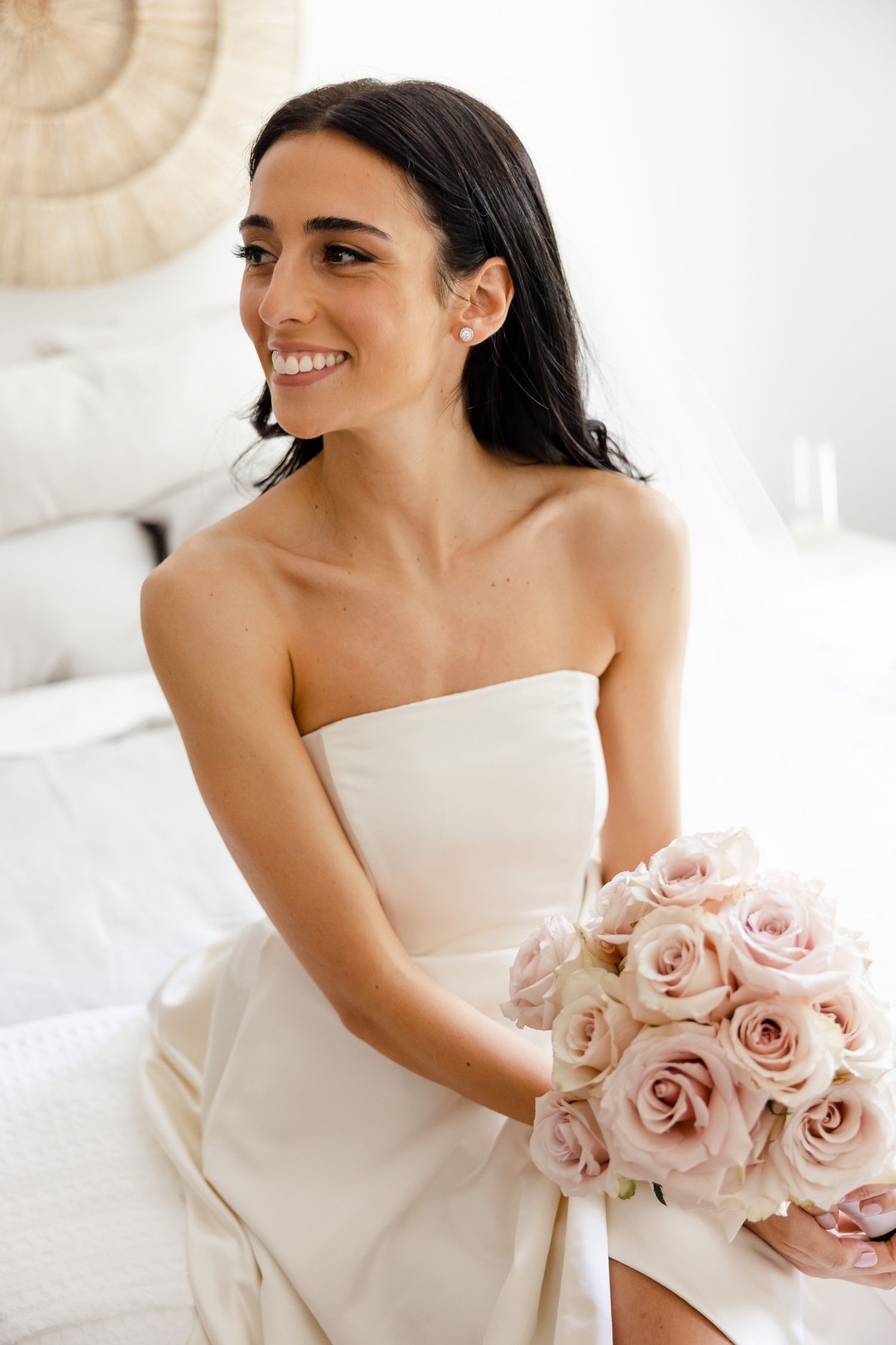 Bride holding pink rose bouquet smiles in elegant strapless wedding gown.