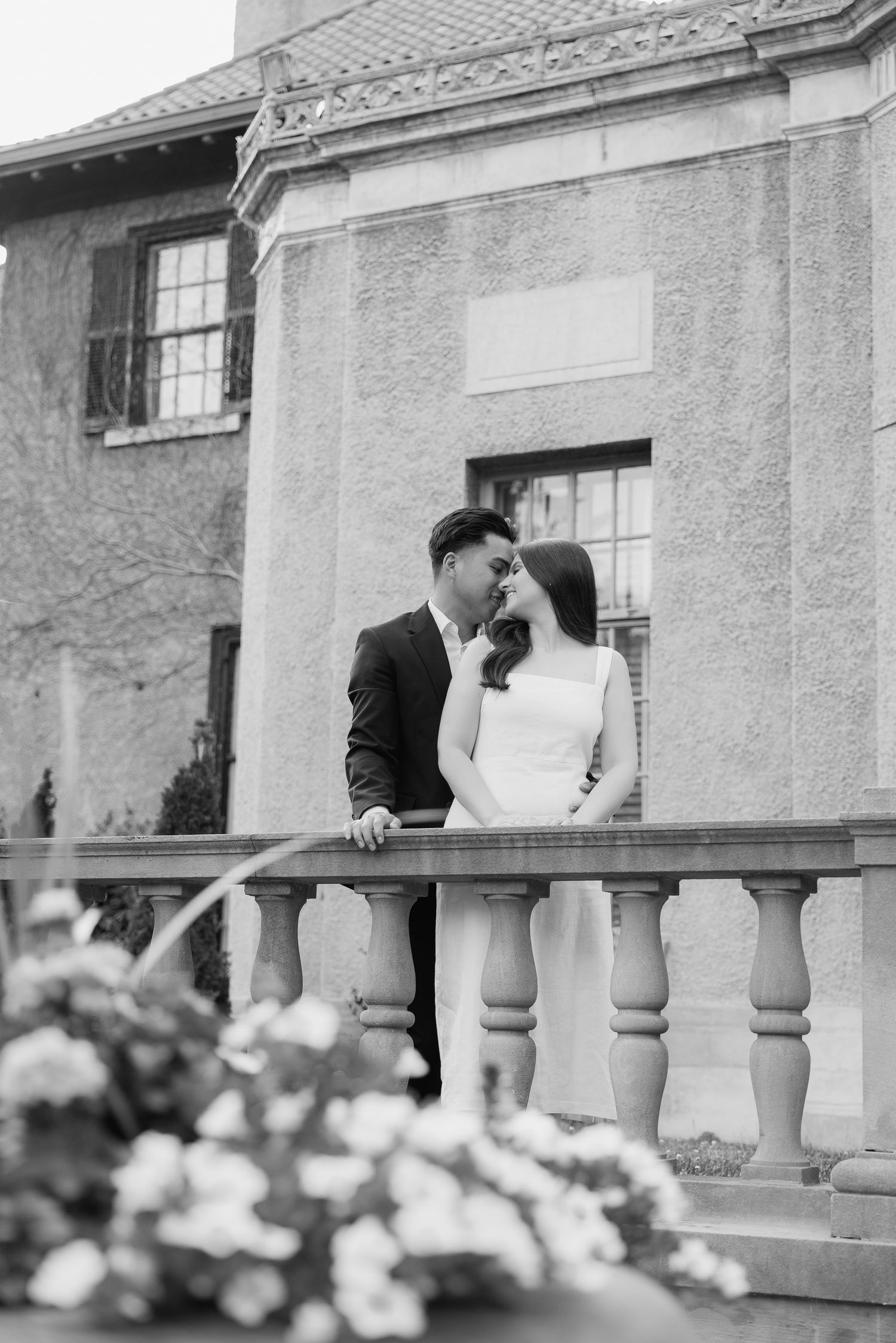 Black and white portrait of couple at ornate stone balustrade with flowers in foreground.