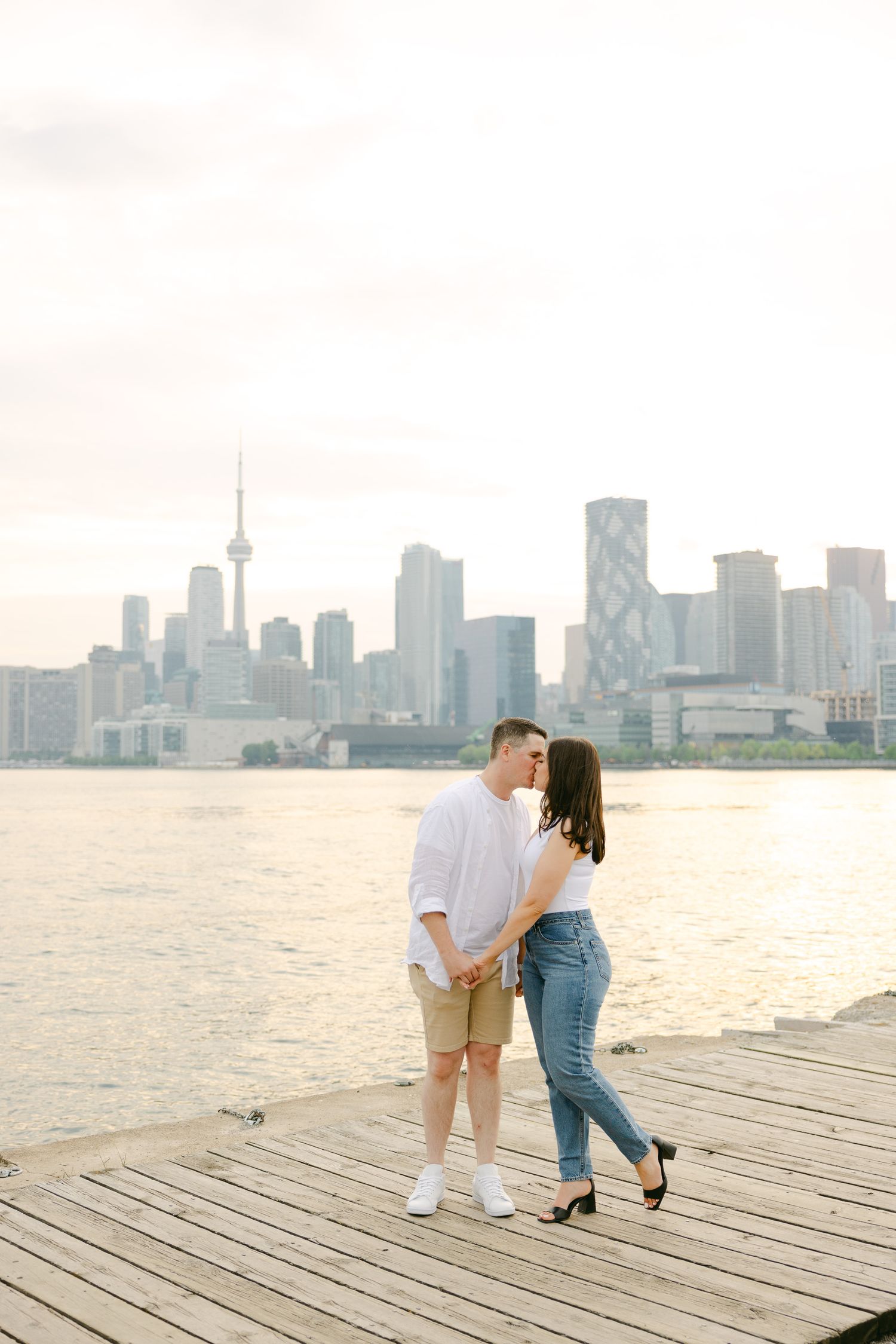 Romantic couple sharing a tender moment on a wooden pier with Toronto's iconic skyline in the background at sunset.