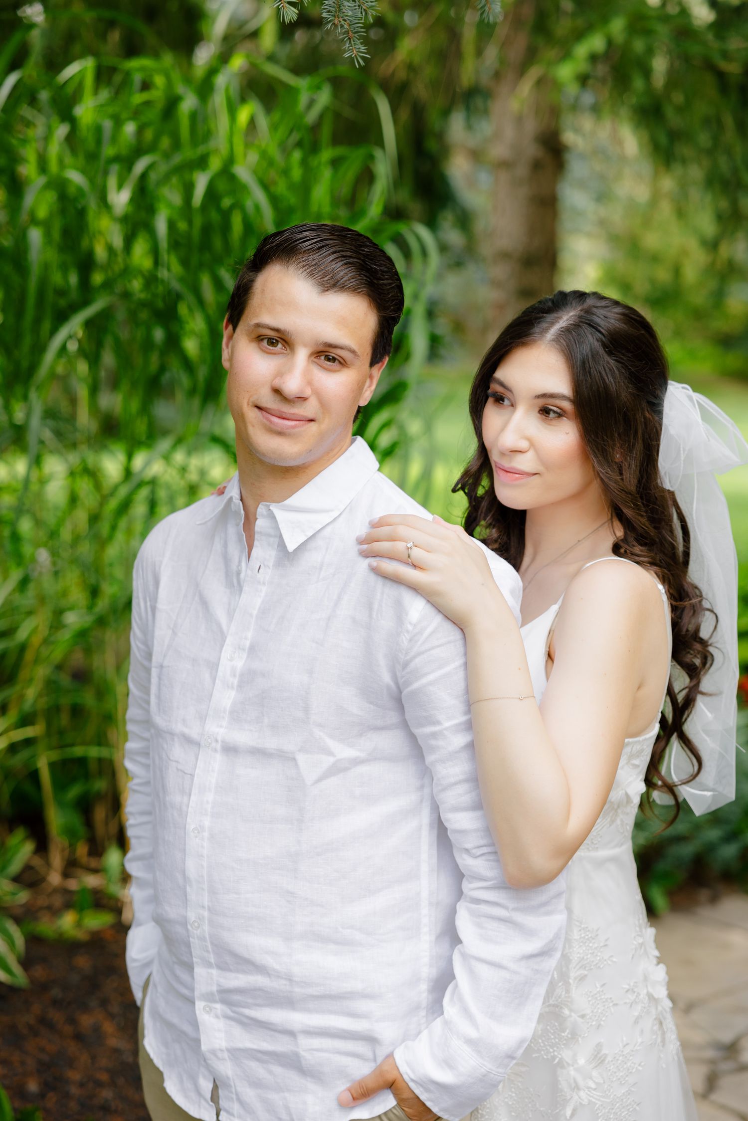 A romantic wedding photo of a couple in white against greenery in a garden setting.