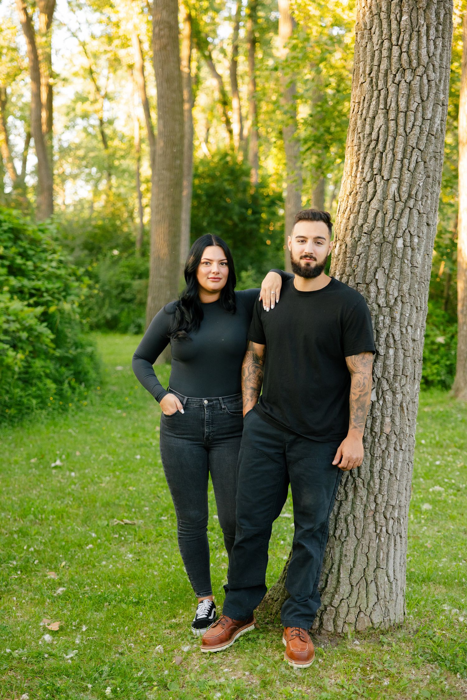 Couple in matching black outfits pose casually against large tree trunk in woodland setting.