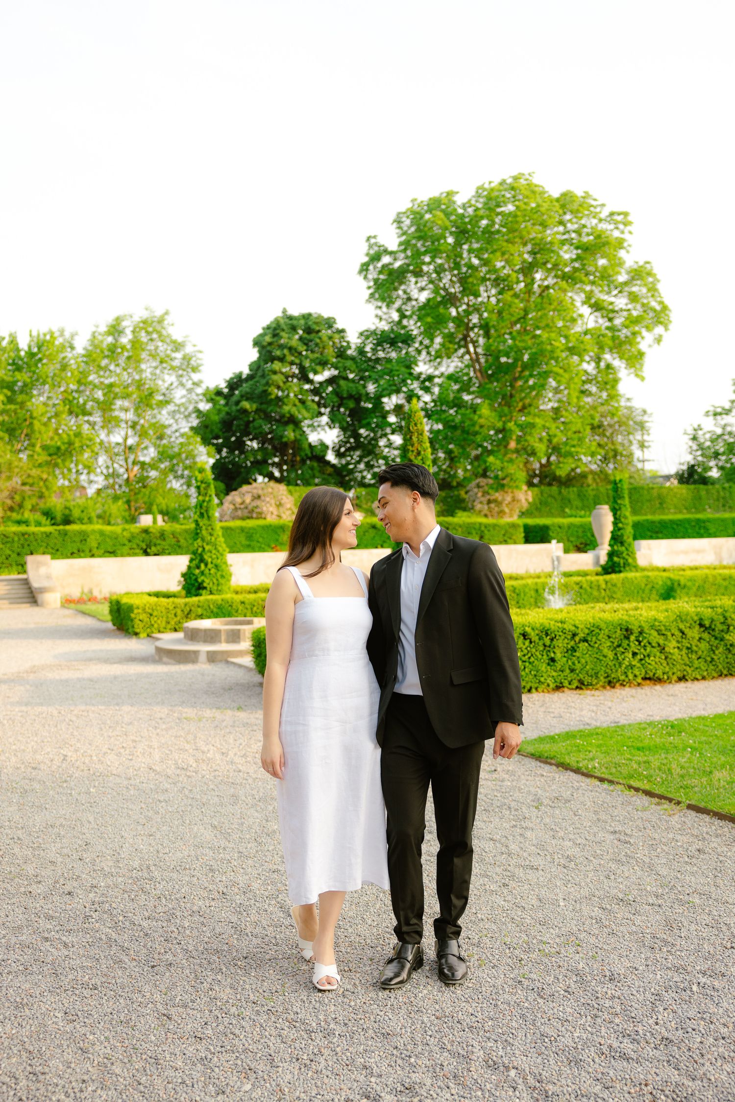 Couple in formal attire poses together on a garden path surrounded by perfectly manicured hedges and trees.