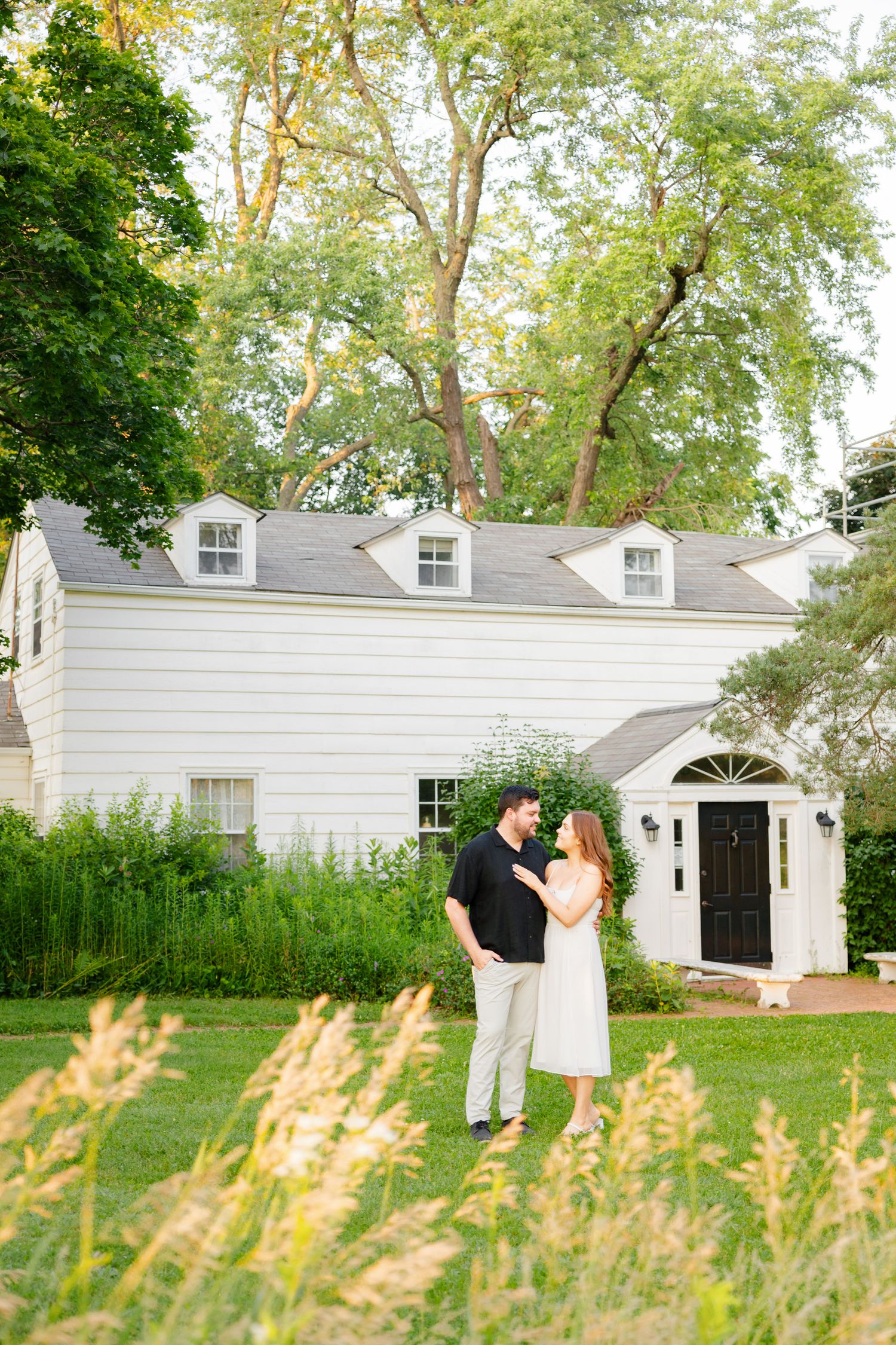 Couple poses romantically in front of classic white house with dormers.