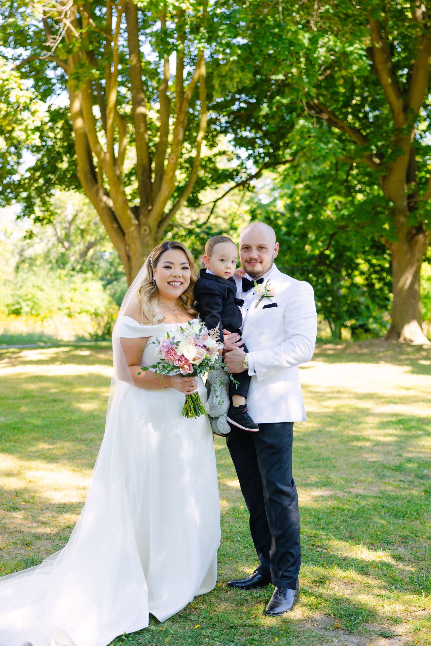 Wedding photo taken outdoors under green trees with a beautiful bride in white dress holding pink bouquet.