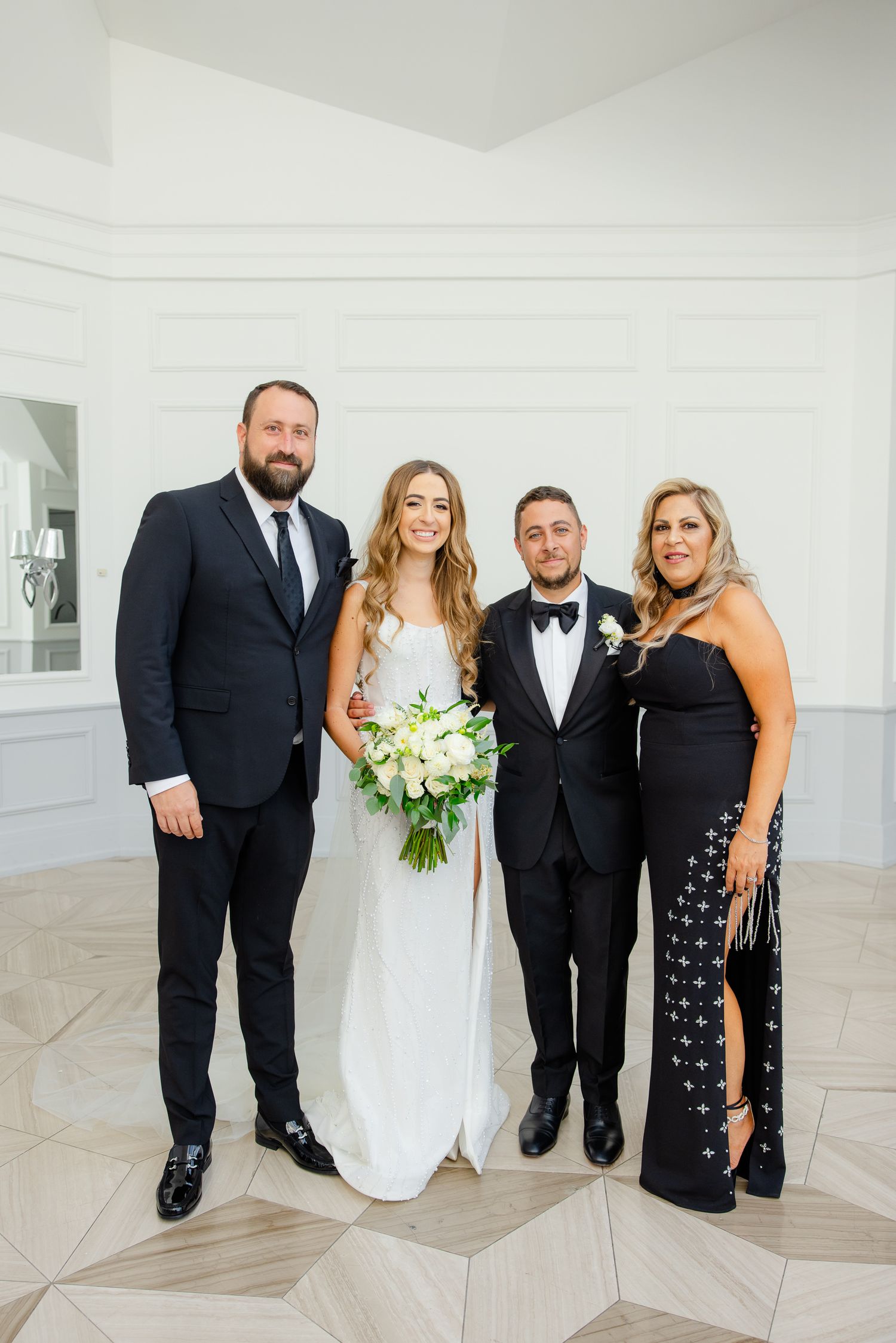 Formal wedding group photo of four people in black tie attire against white interior setting.