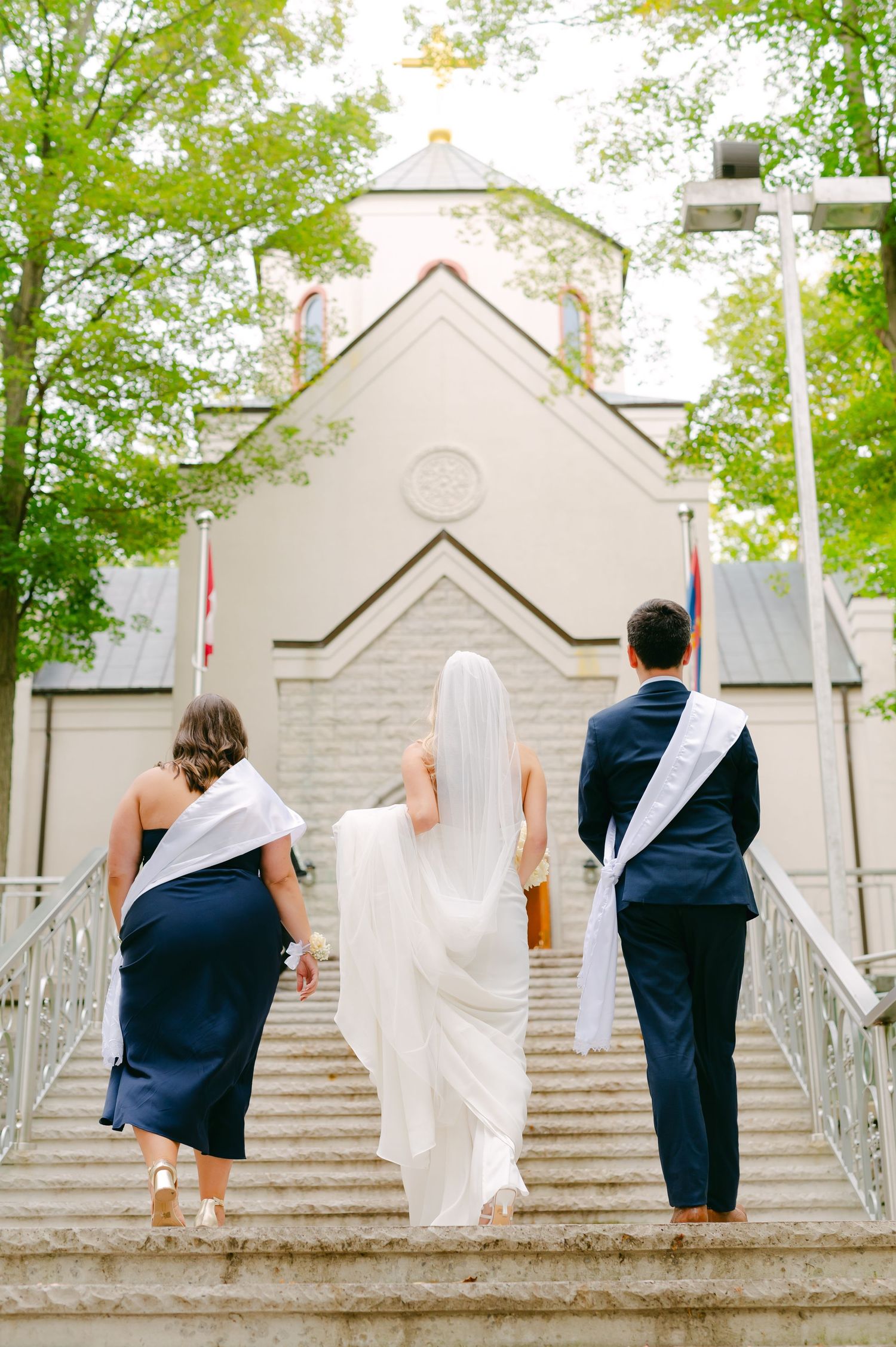Three people ascending church steps with flowing white wedding dress in center.
