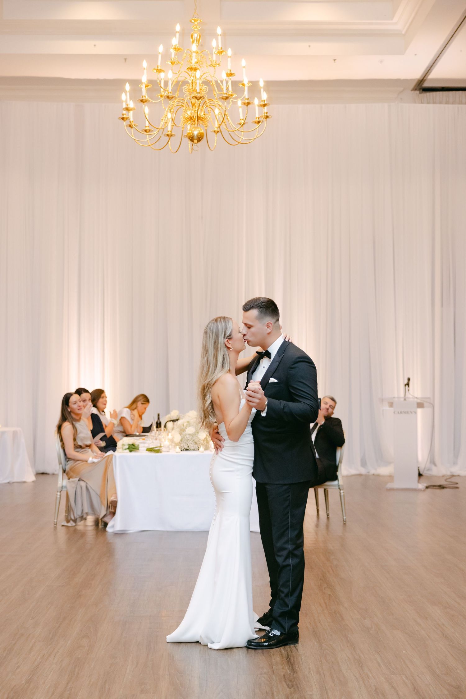 A couple dances in an elegant ballroom with white draping and a gold chandelier overhead.