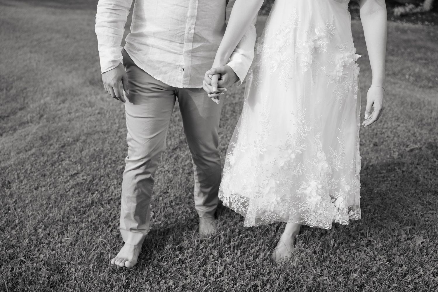 Black and white photo showing barefoot legs of a couple on grass, bride in lace dress and groom in khakis.