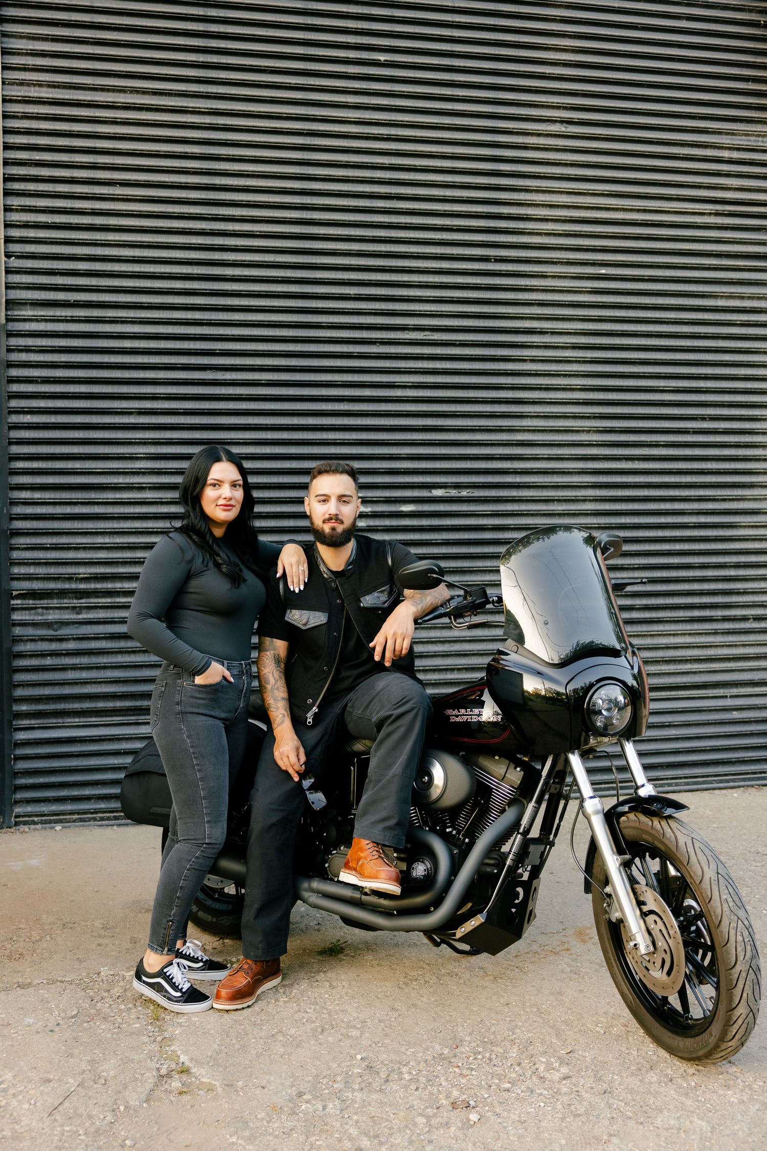 A couple in dark clothing poses together with their custom Harley Davidson motorcycle.