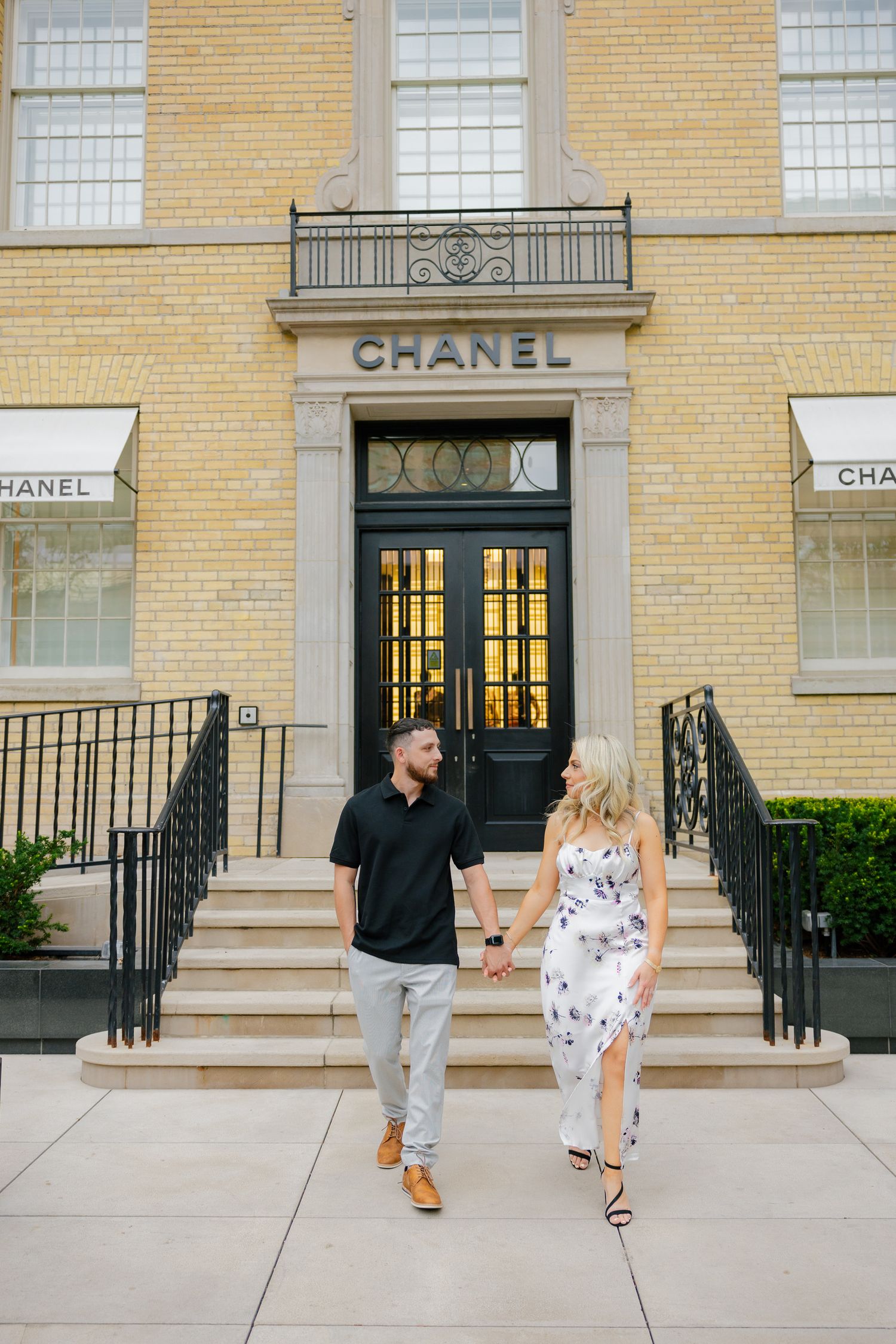 A couple walks down steps in front of the Chanel store with yellow brick facade and elegant railings.